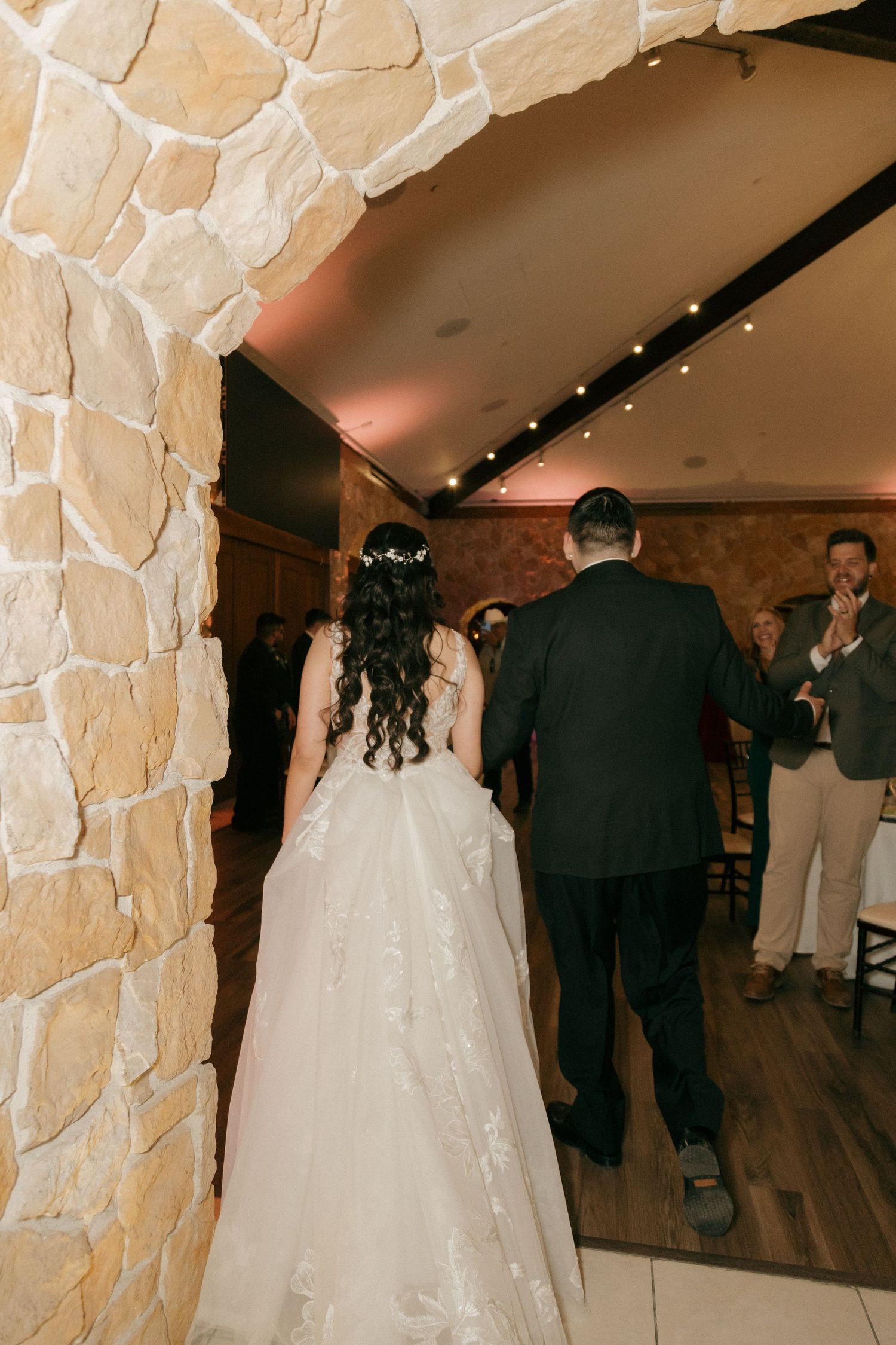 Bride in white ballgown enters stone-walled wedding reception hall with escort as guests applaud.