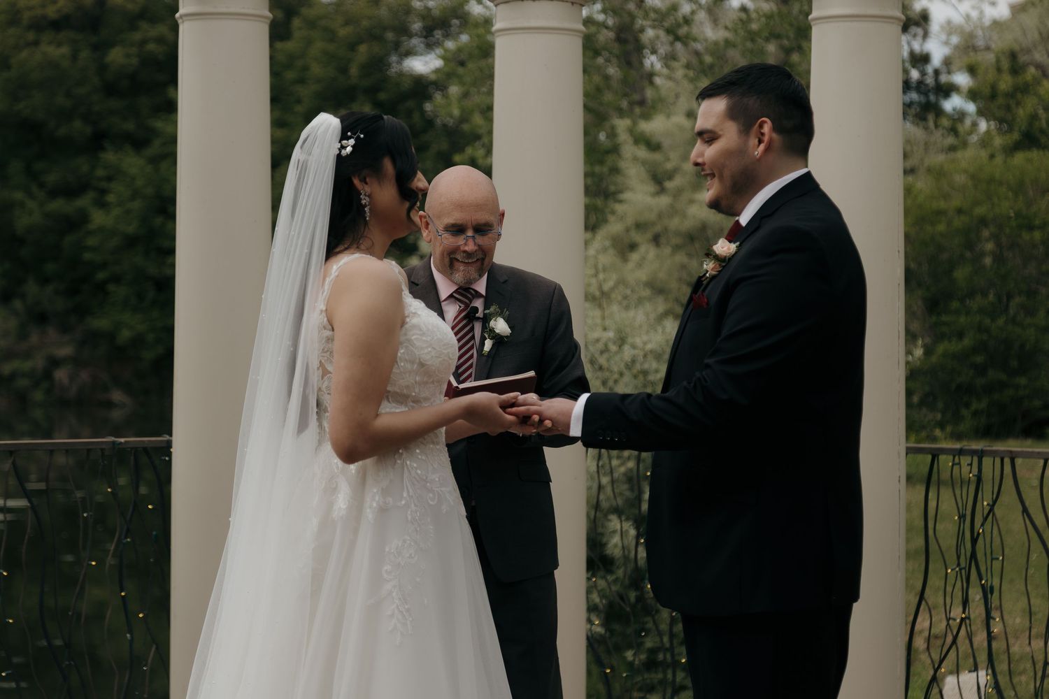 Bride and groom exchange rings during outdoor wedding ceremony with white columns and lush greenery backdrop.