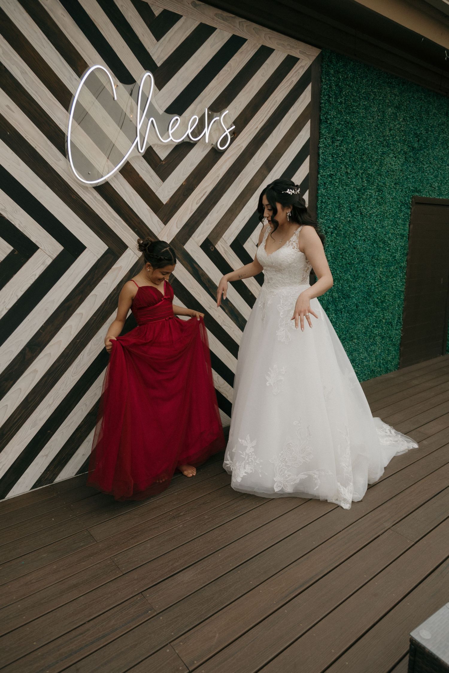Bride in white gown and bridesmaid in red dress pose together near striped wall with Cheers text.