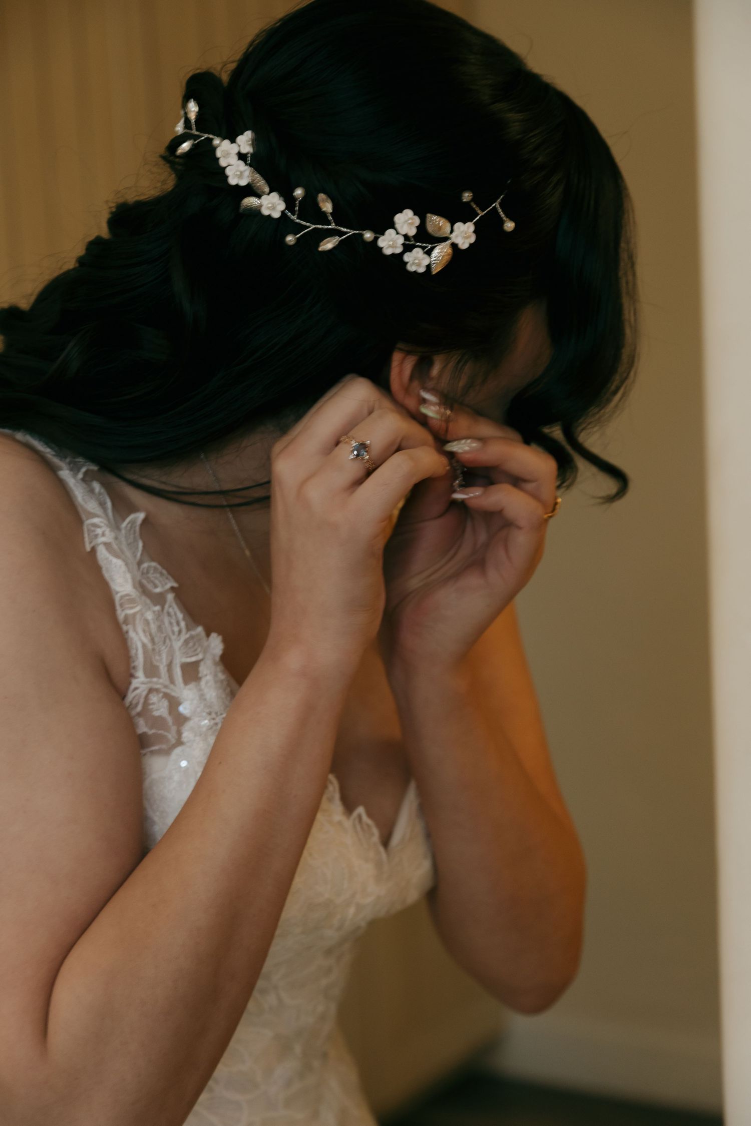 Bride with floral hair vine putting on earrings in white lace wedding gown.