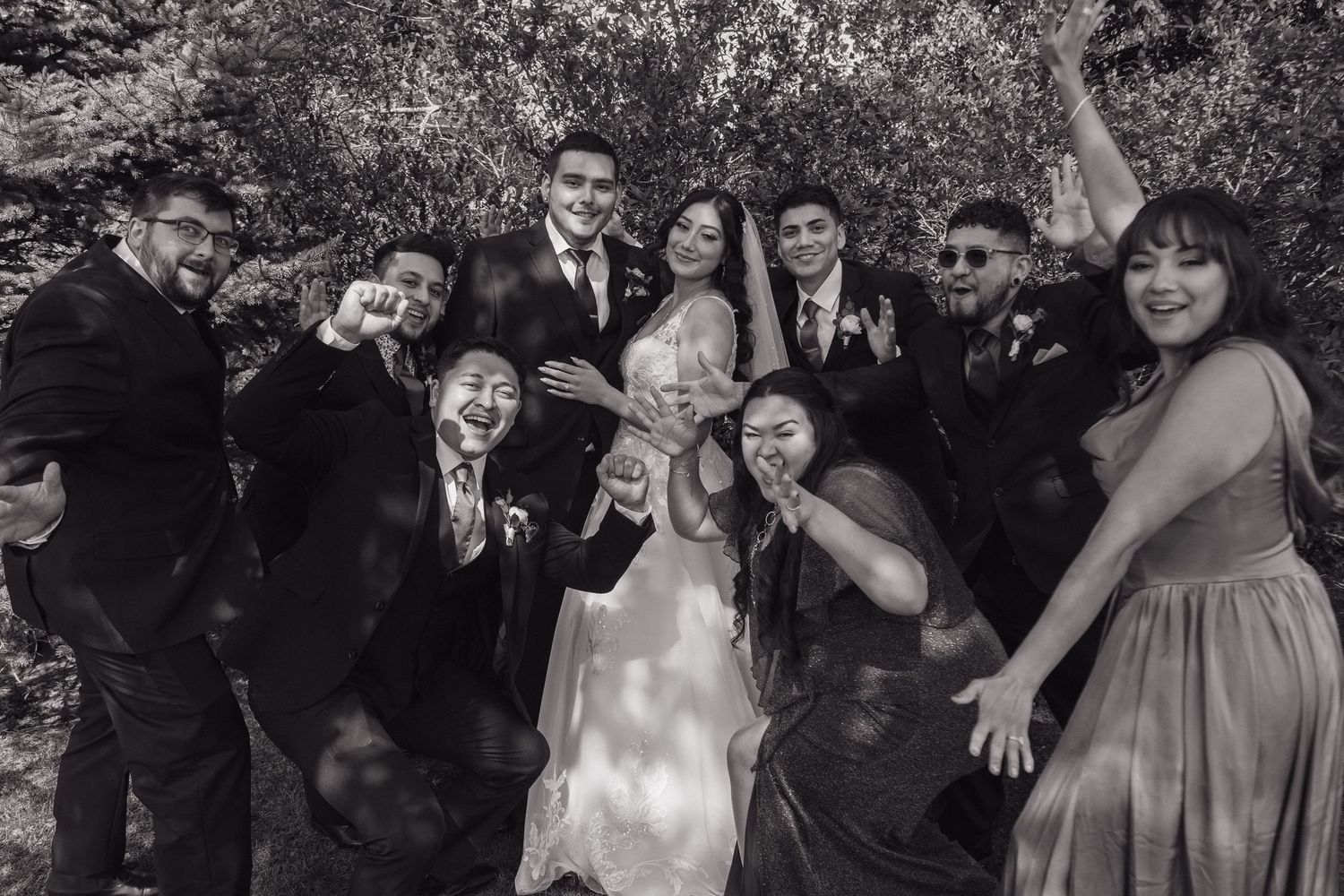 Black and white fun group photo of wedding party celebrating with lively poses under leafy trees.