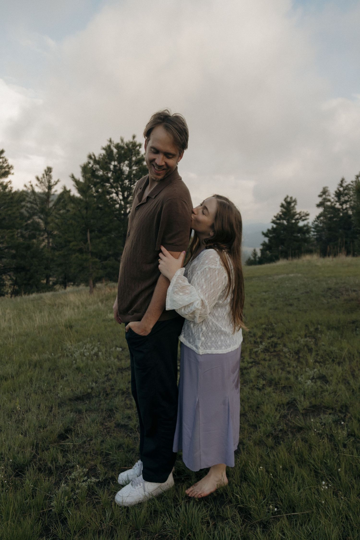 Couple embracing on a green hillside, woman kissing mans cheek while he smiles outdoors.