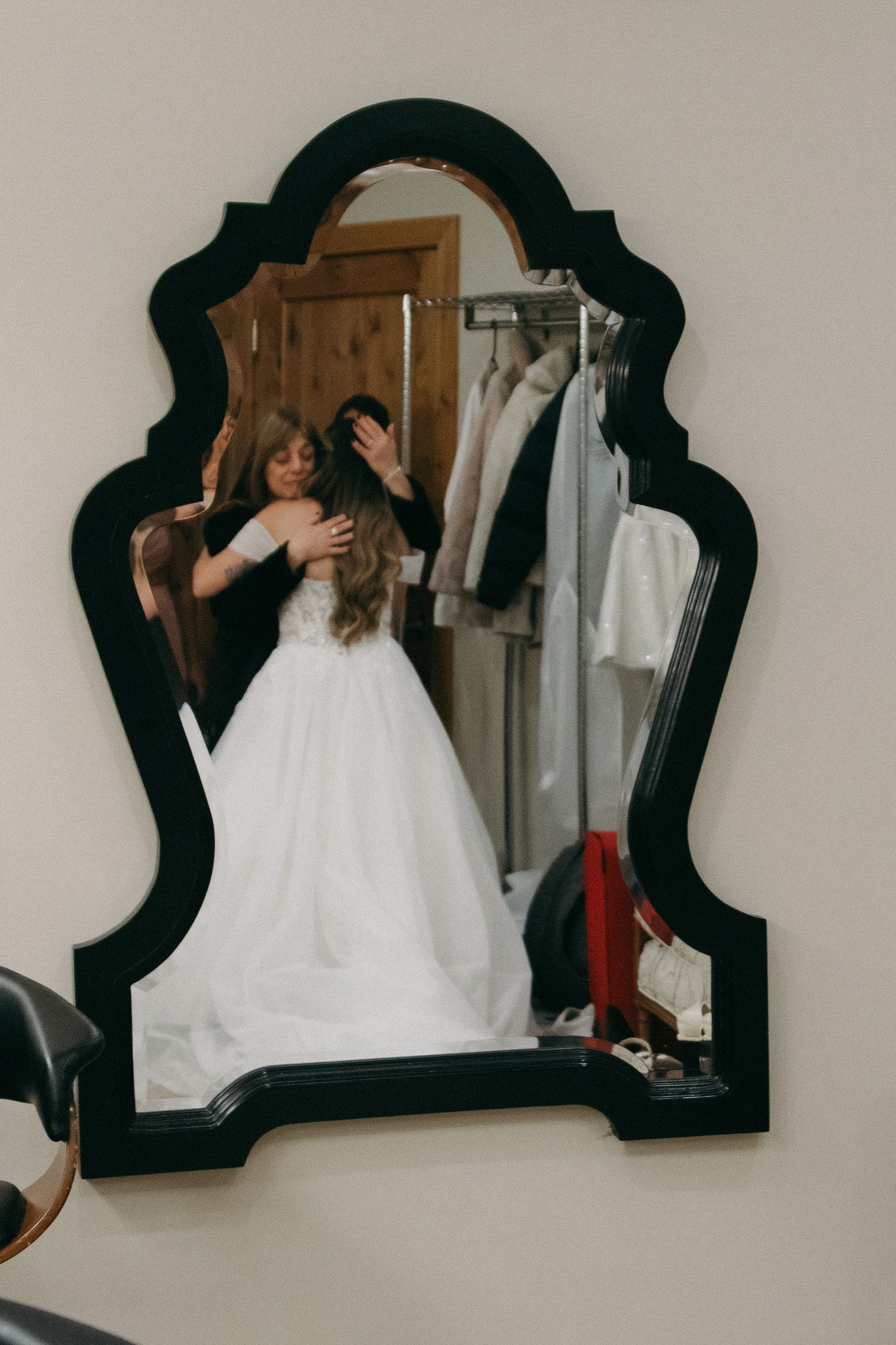 Bride trying on white lace wedding gown reflected in decorative mirror.