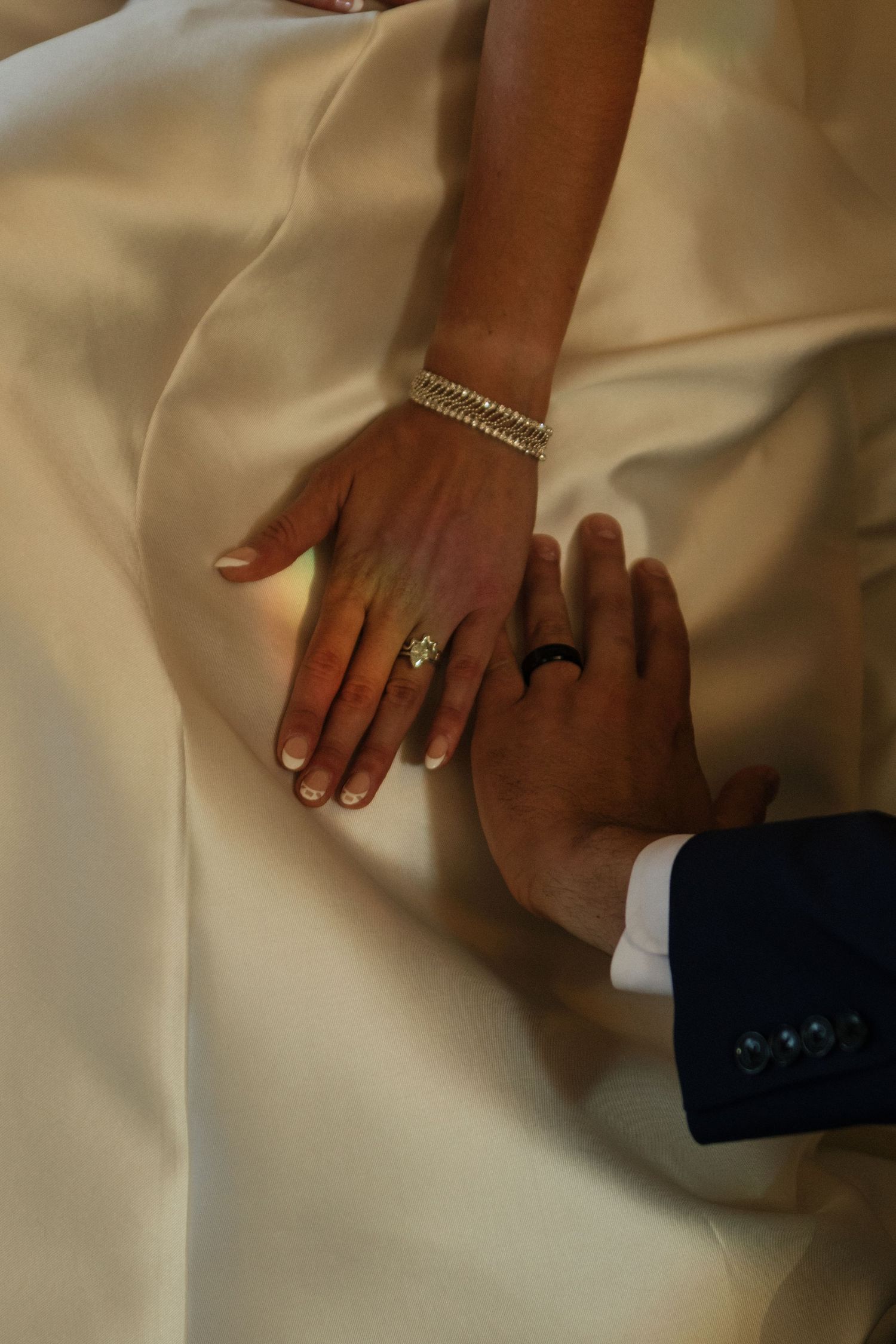 Wedding rings and hands photographed in soft lighting against white fabric creating an intimate moment.