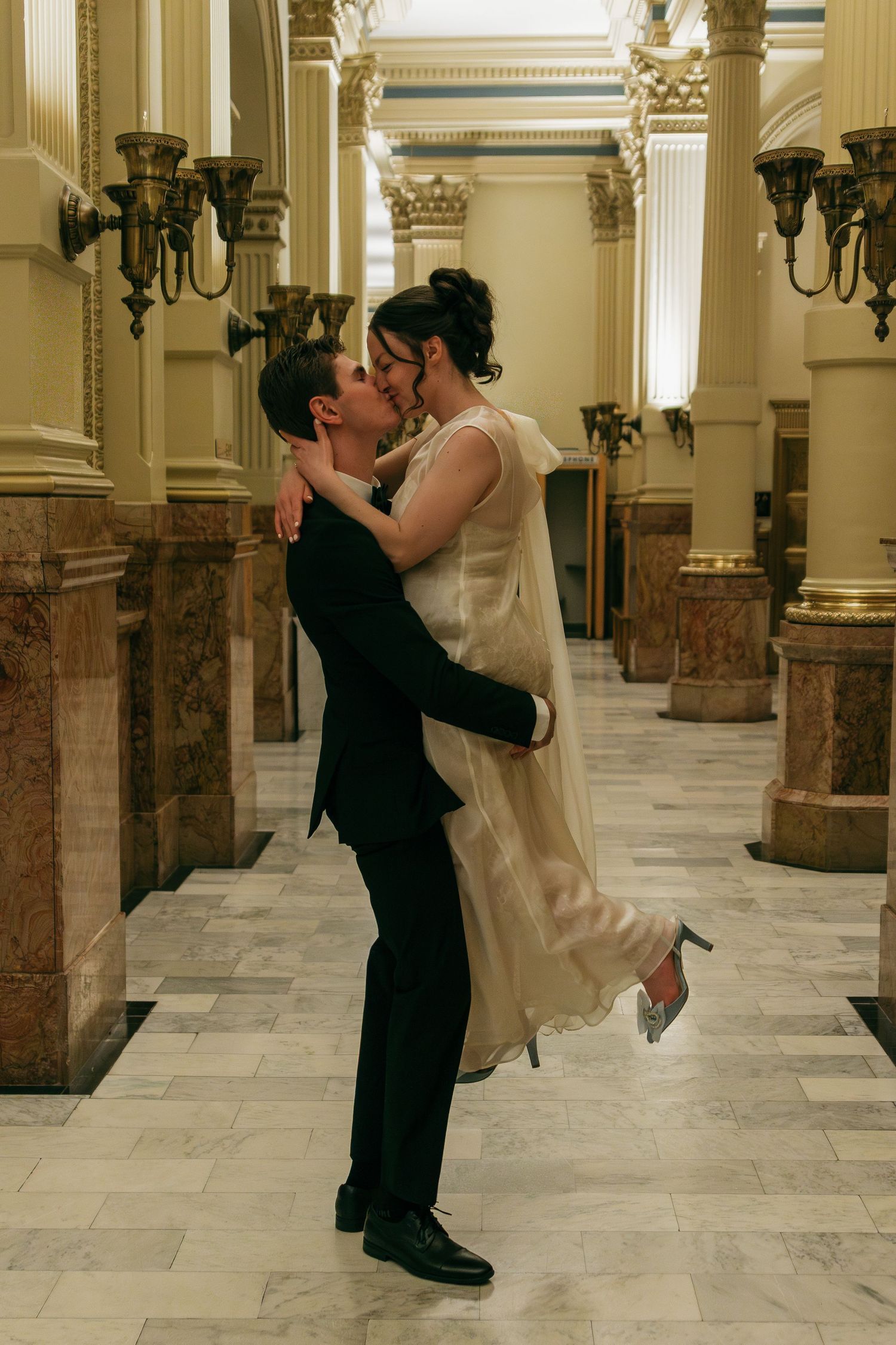 Wedding couple performing dramatic dance dip in marble hallway with columns.