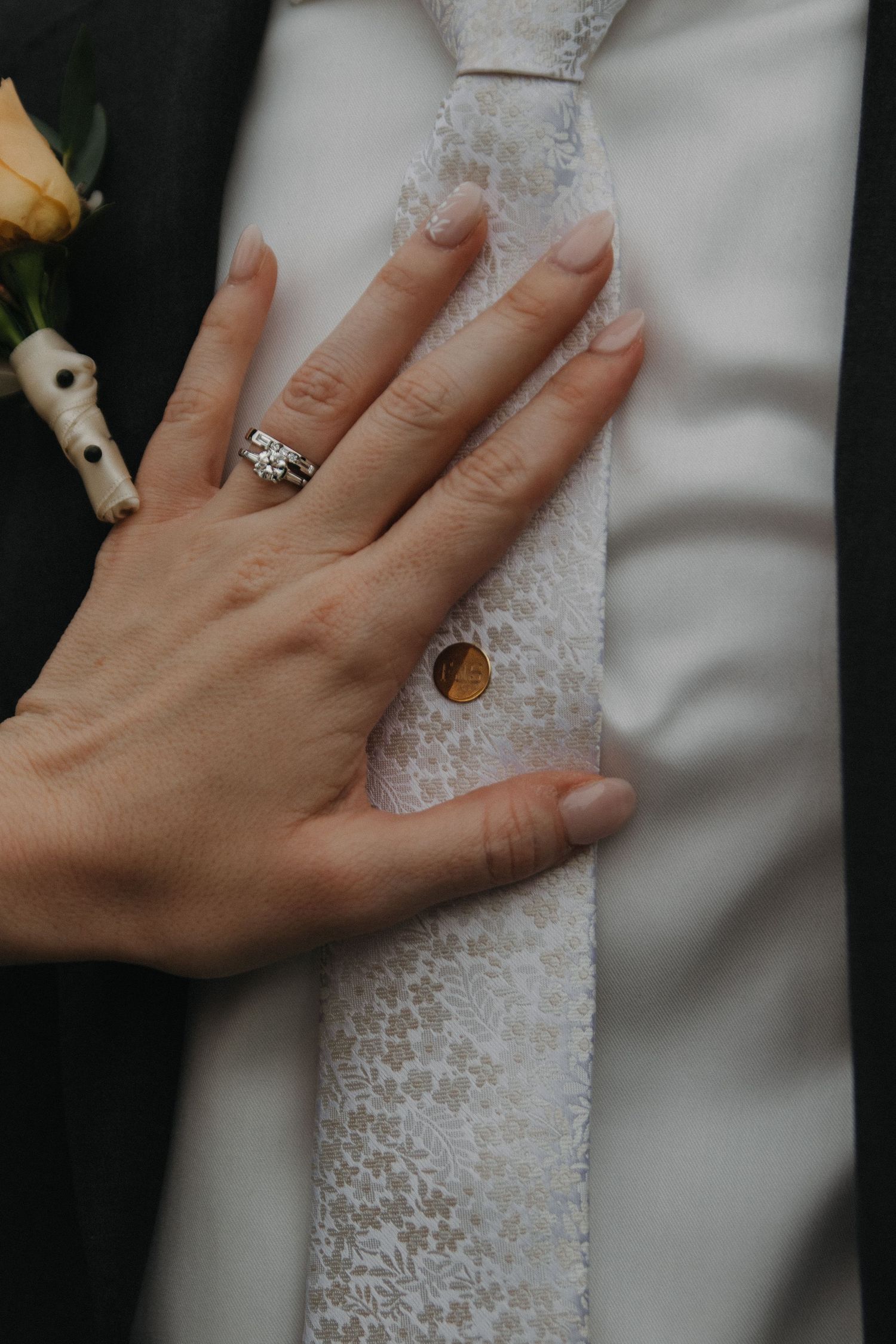 A hand wearing an elegant engagement ring rests against white lace fabric with delicate embroidery details.