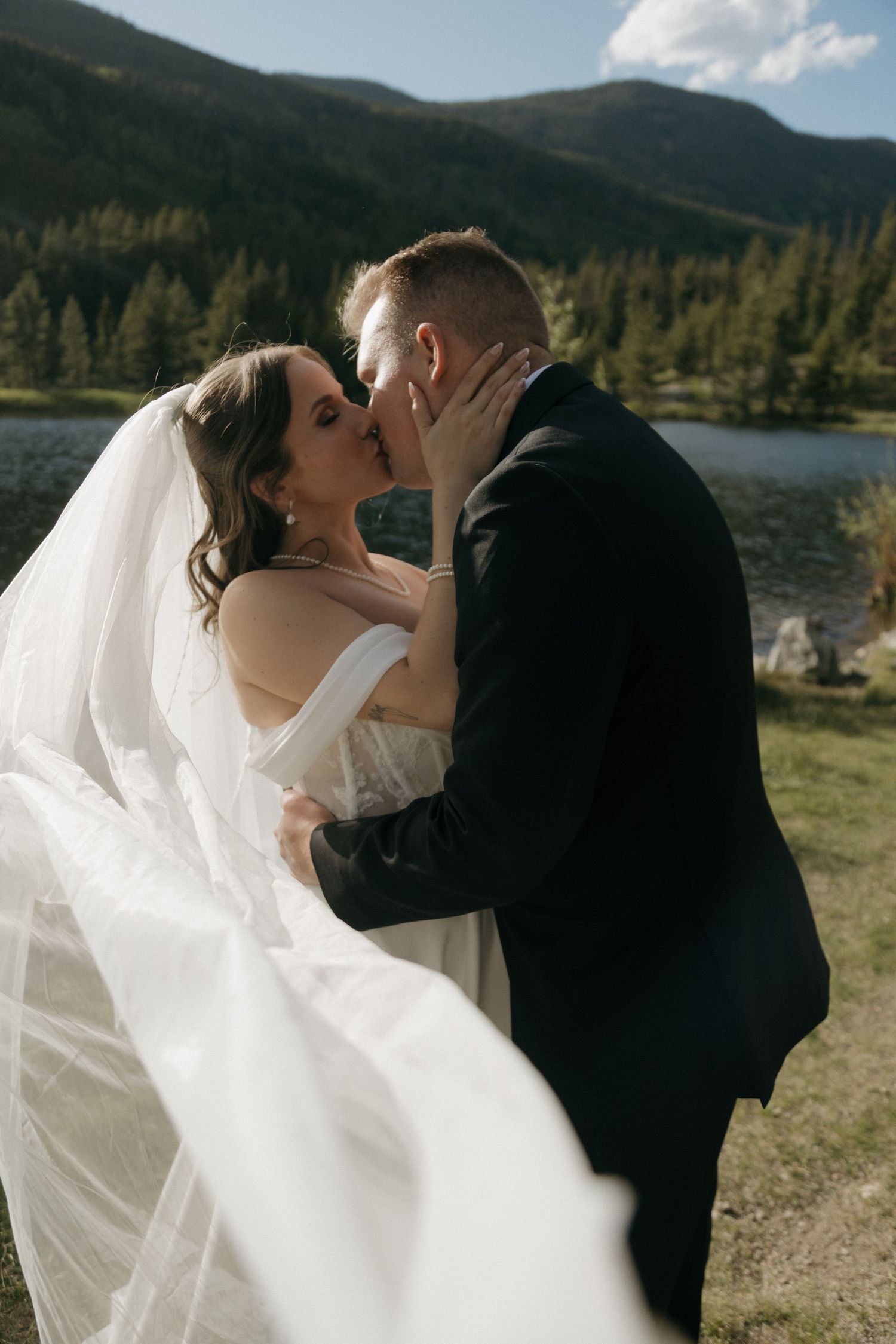 Bride and groom share a romantic kiss outdoors with a flowing veil, mountain lake and pine forest backdrop.