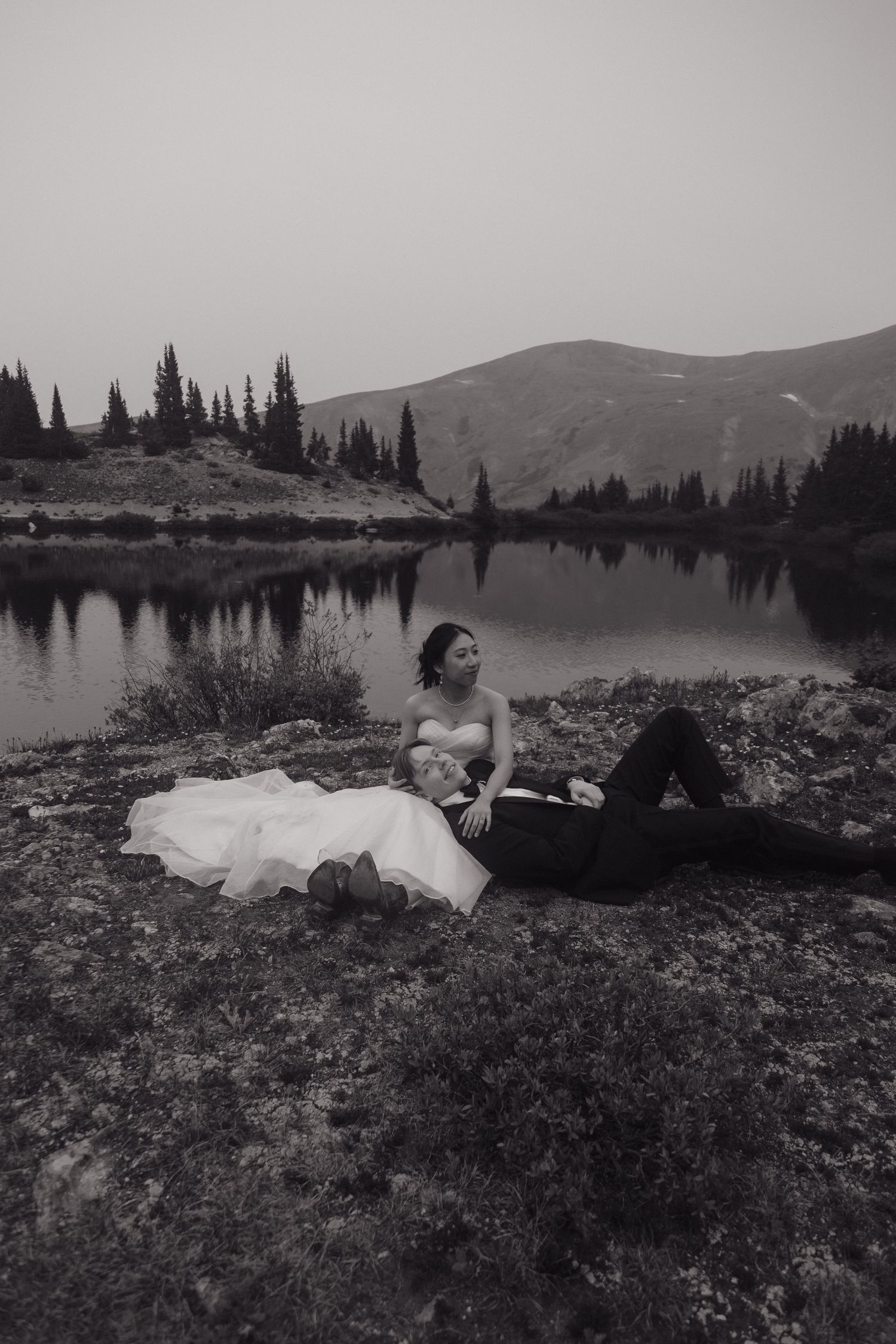 Black and white photo of bride and groom relaxing by a serene alpine lake with mountain reflections.