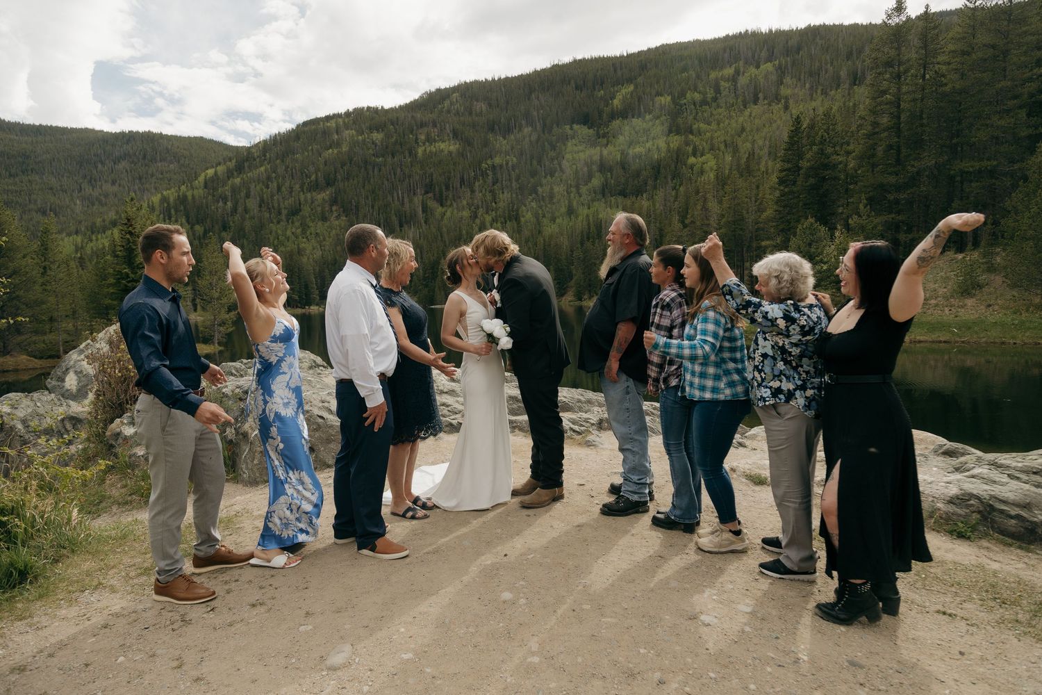 Wedding party celebrating outdoors by a mountain lake with forested hills in background.