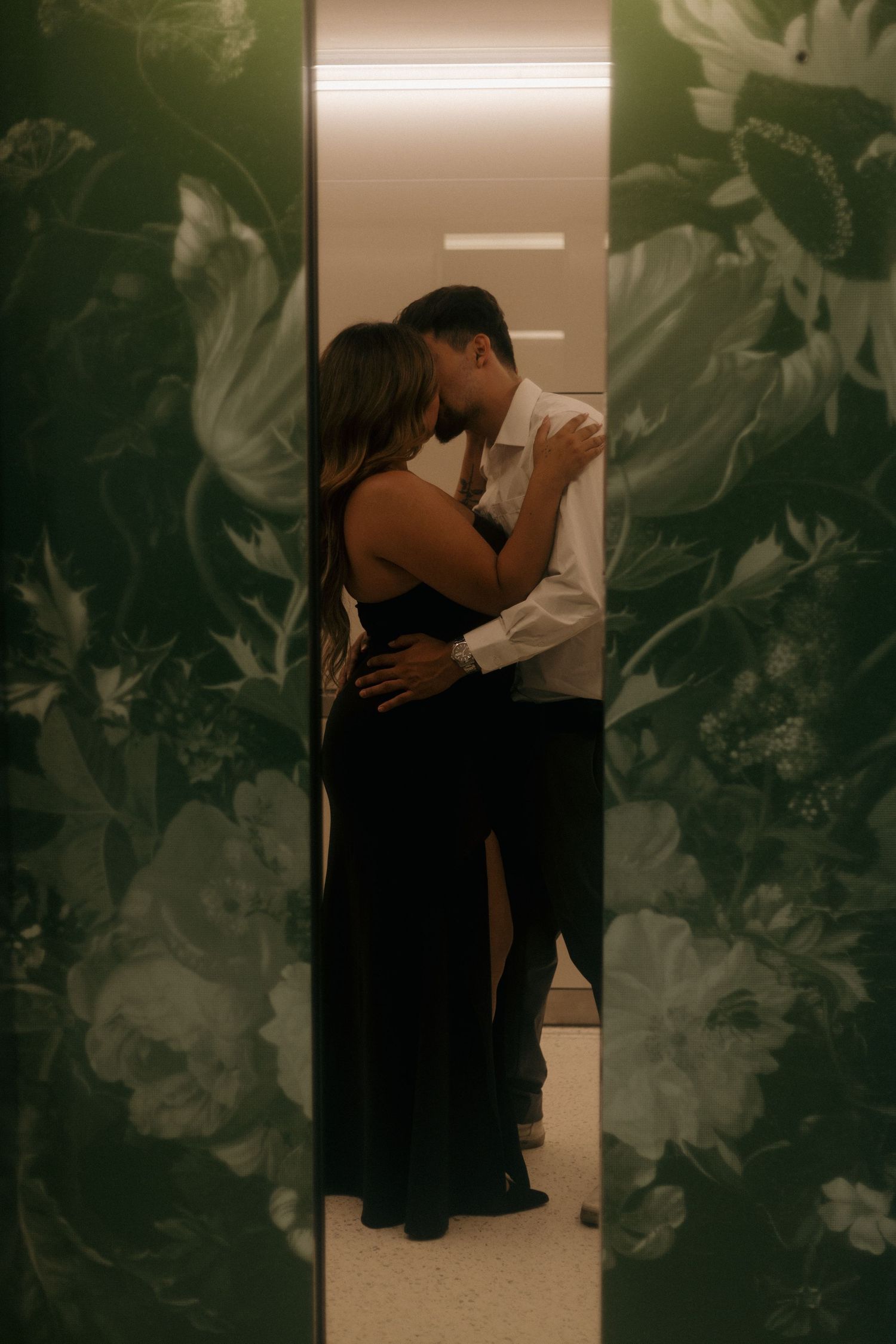 Couple embracing and kissing in an elevator framed by dark floral wallpaper.
