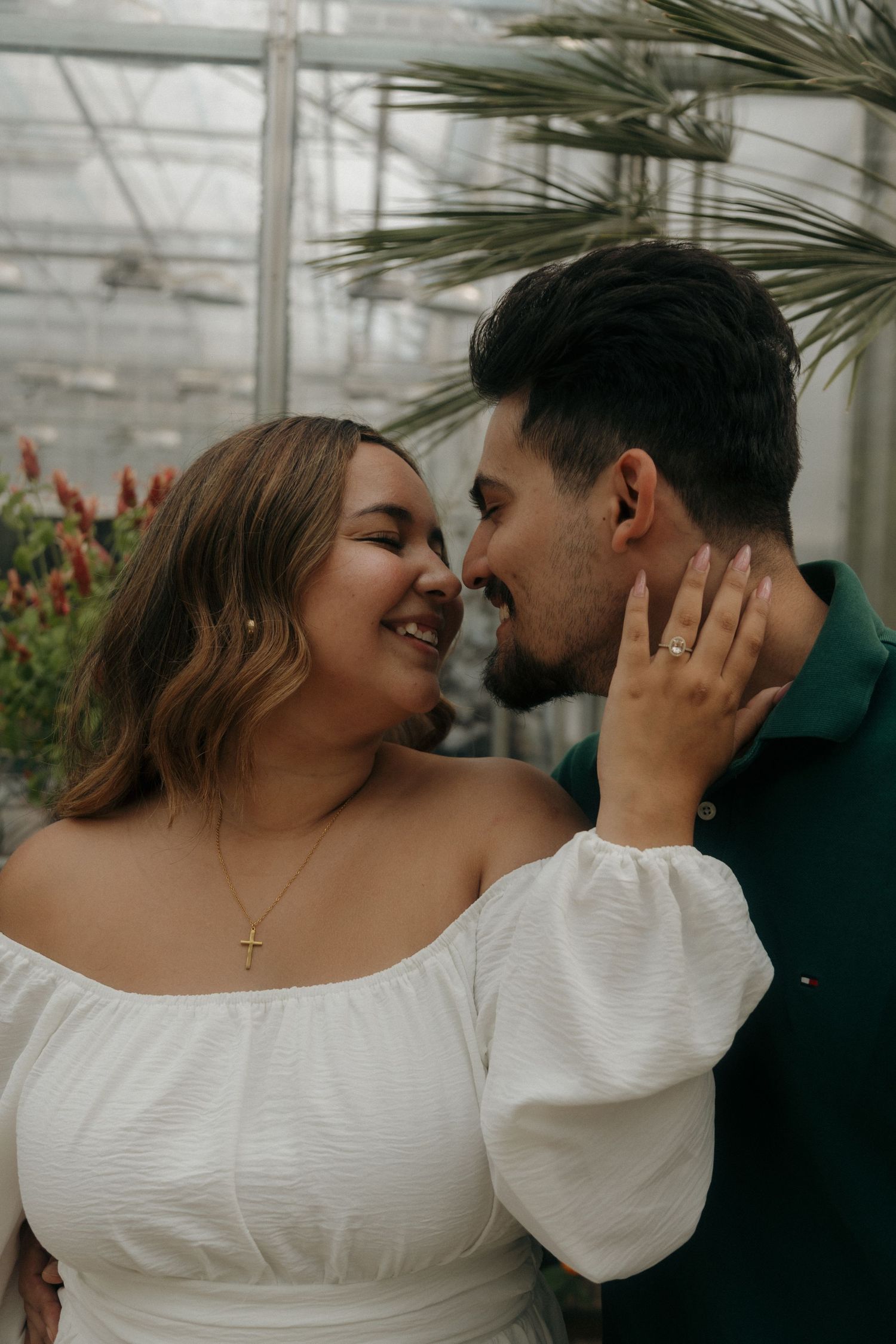 Couple sharing a tender moment in a greenhouse, woman in white off-shoulder dress touching mans face.