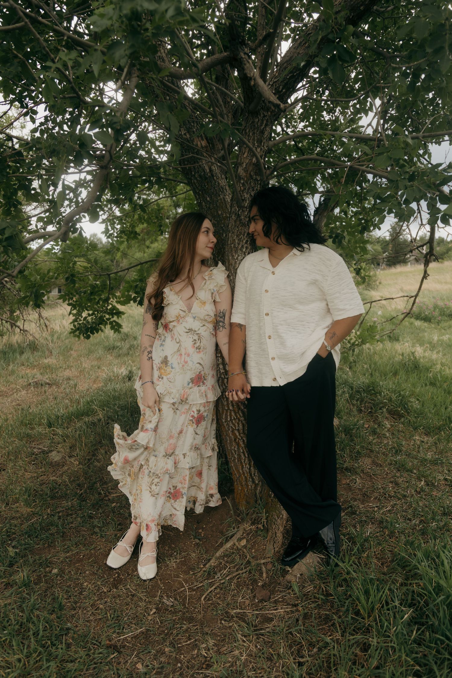 Couple holding hands leaning against tree trunk in lush green field, woman in floral maxi dress and man in white shirt.