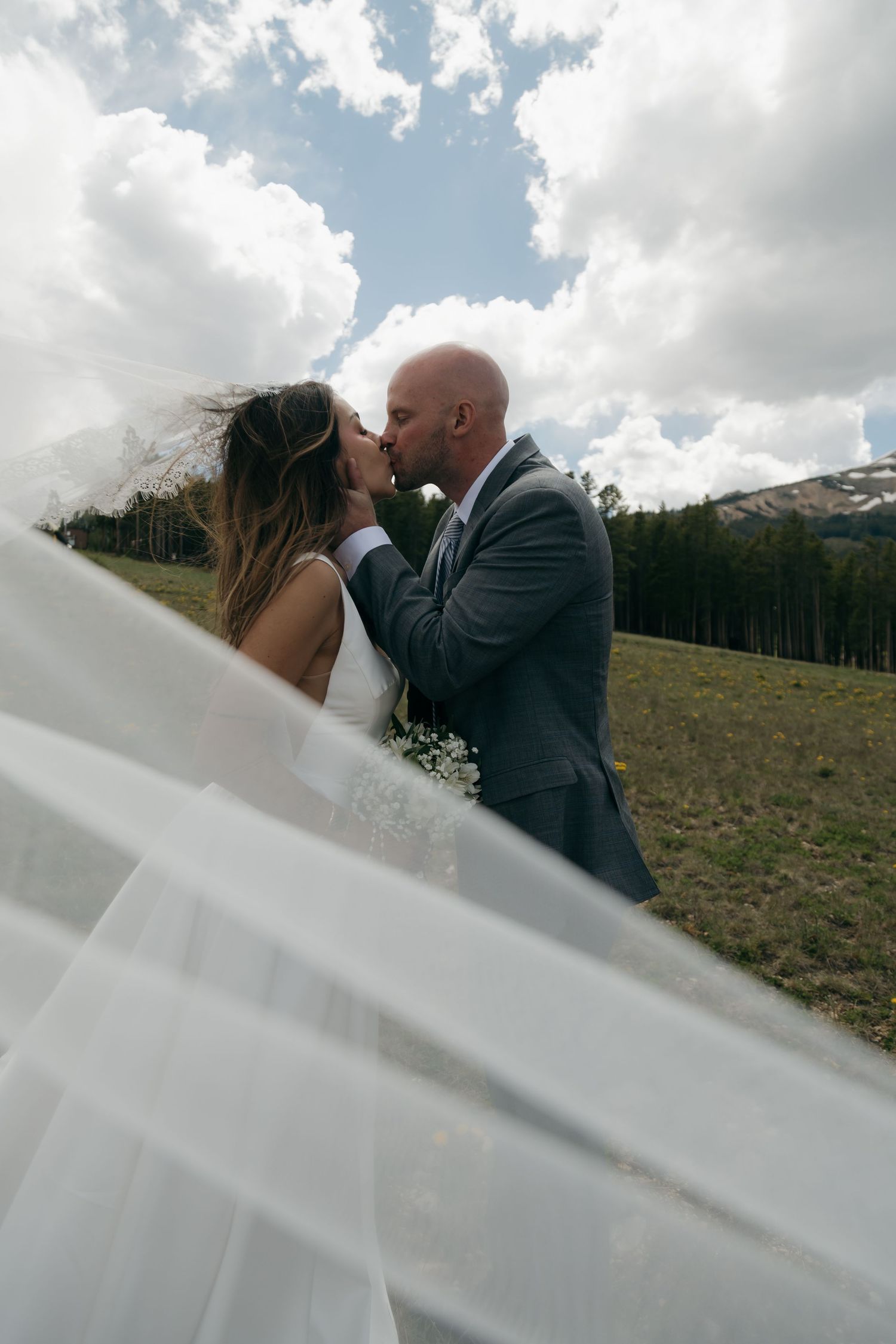 Bride and groom share a kiss in mountain meadow with veil billowing in the wind.