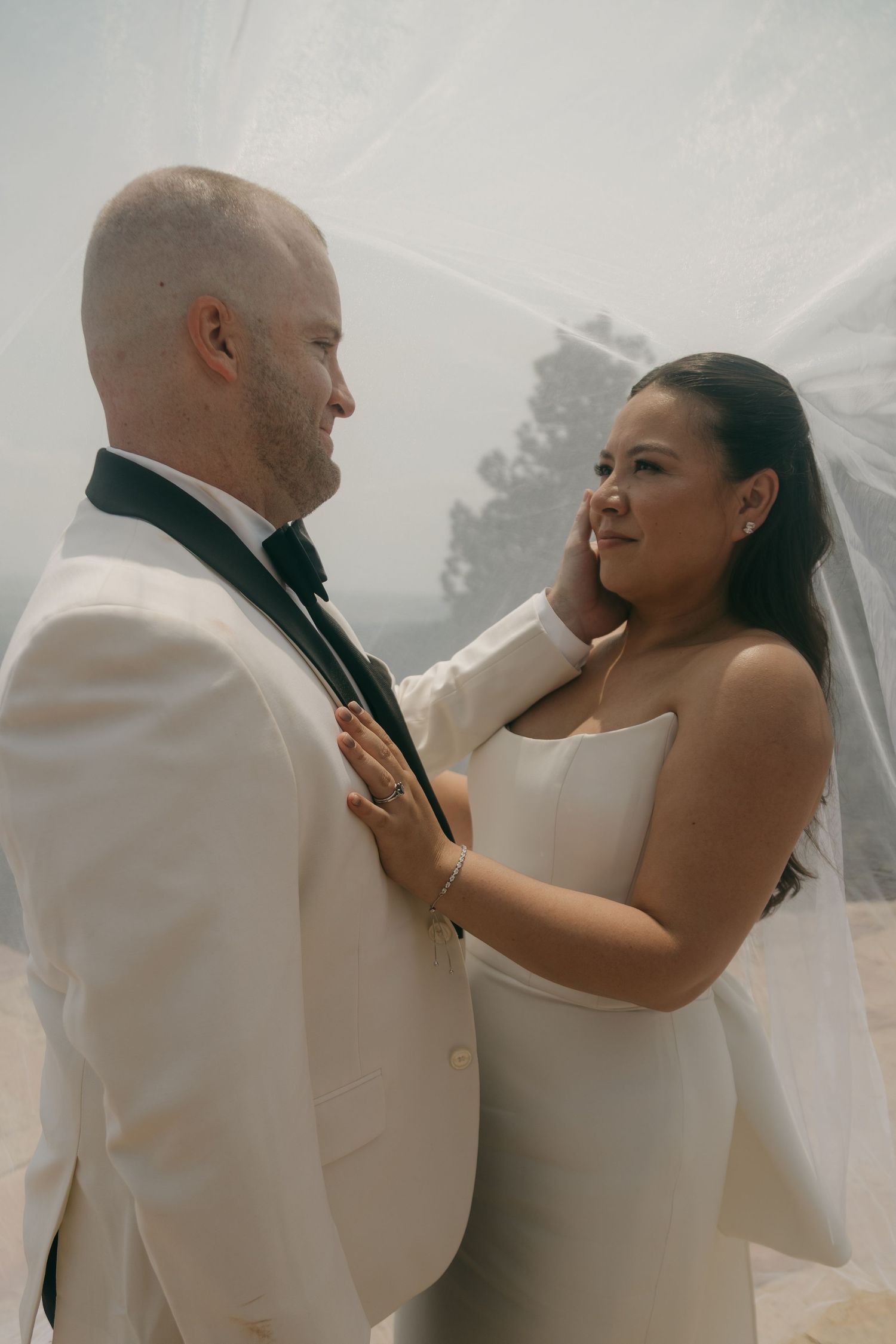 Groom in white tuxedo tenderly cups brides face under veil at outdoor wedding.