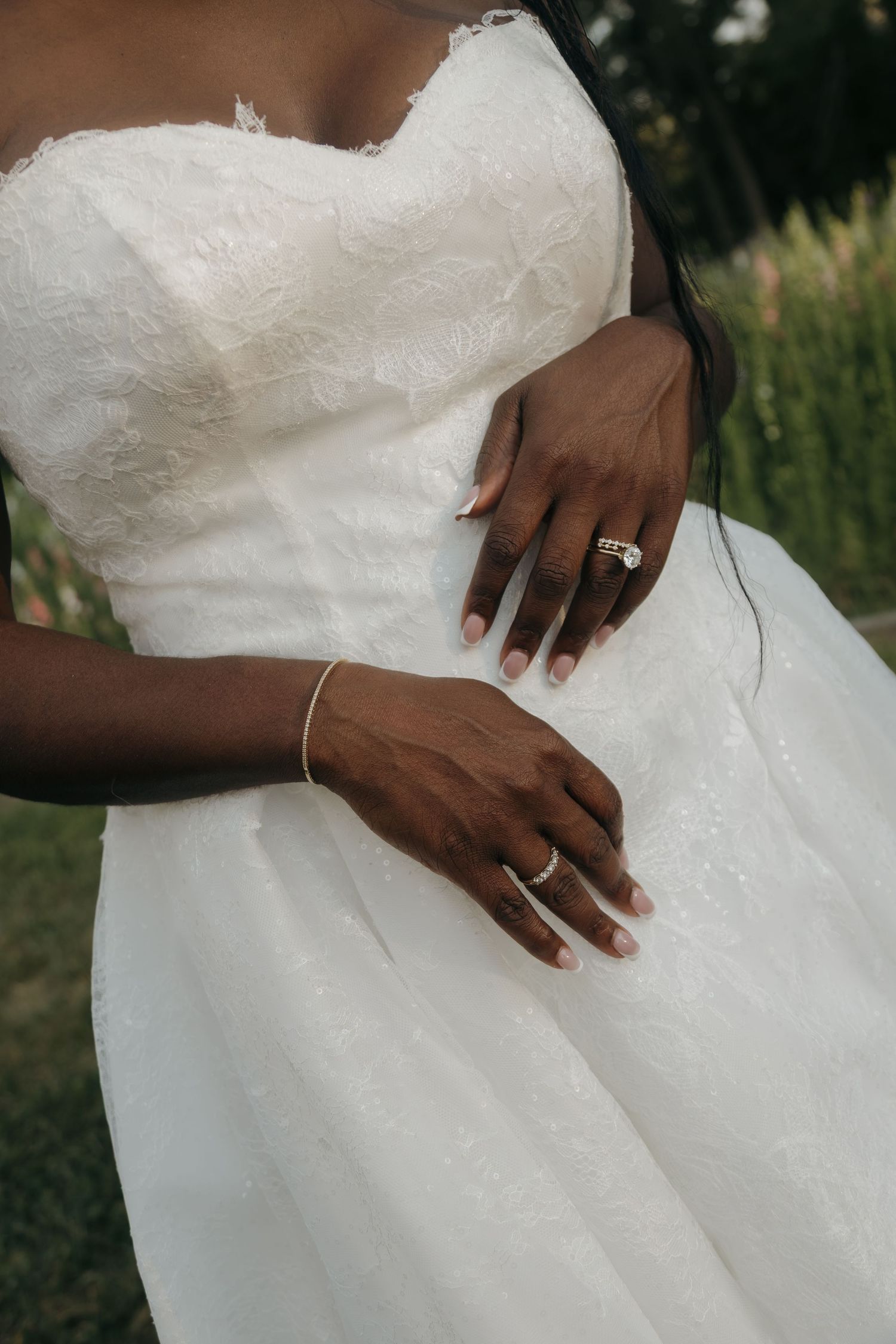 Close-up of a Black bride's hands showing wedding rings and bracelet against her white lace strapless wedding gown.