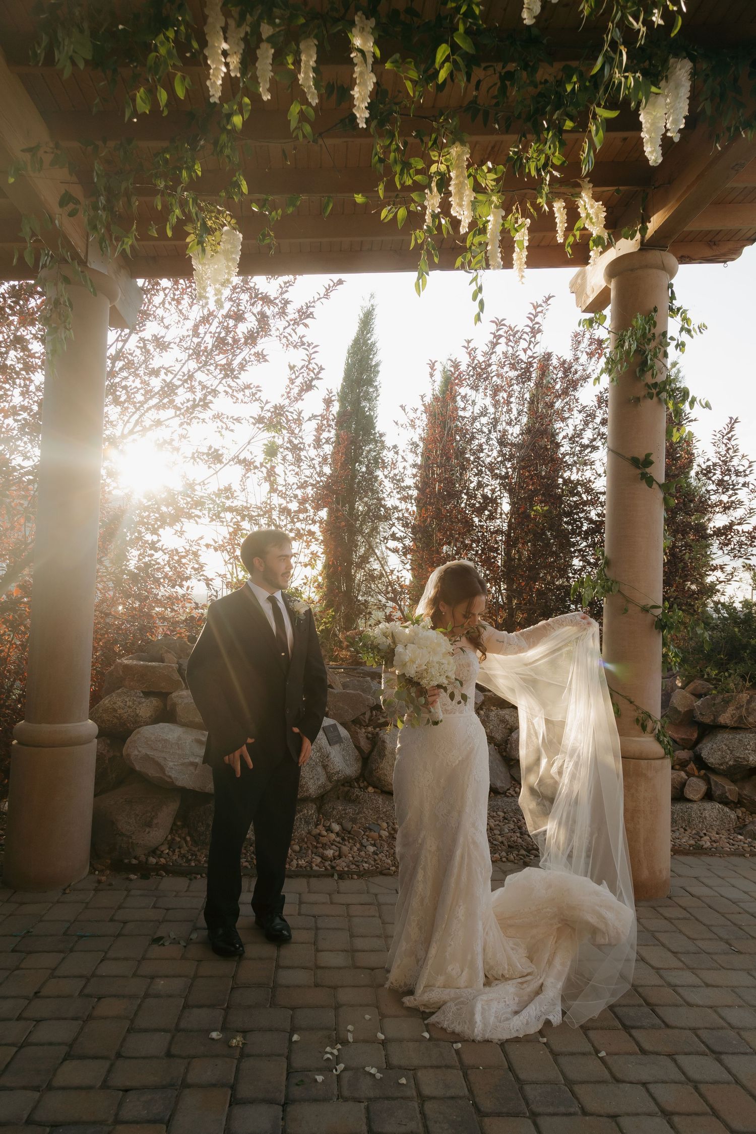 Bride and groom share golden hour moment under pergola with flowing veil and bridal bouquet.
