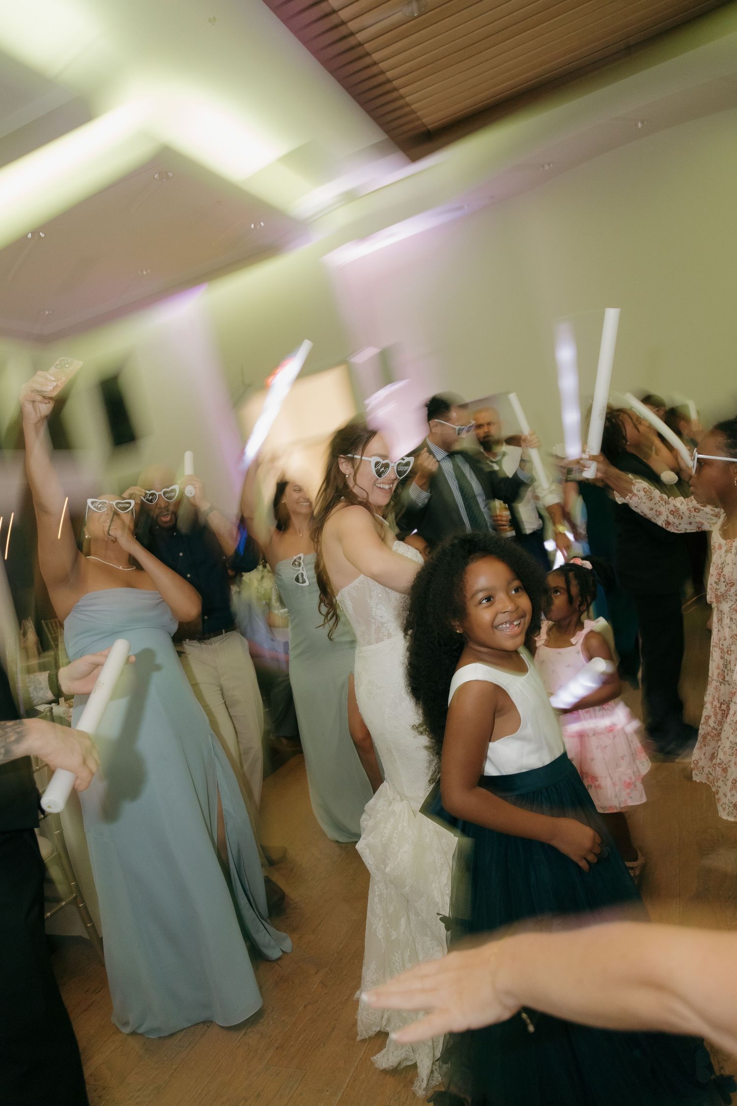 Guests wave glow sticks on dance floor with young girl smiling at lively wedding reception party.