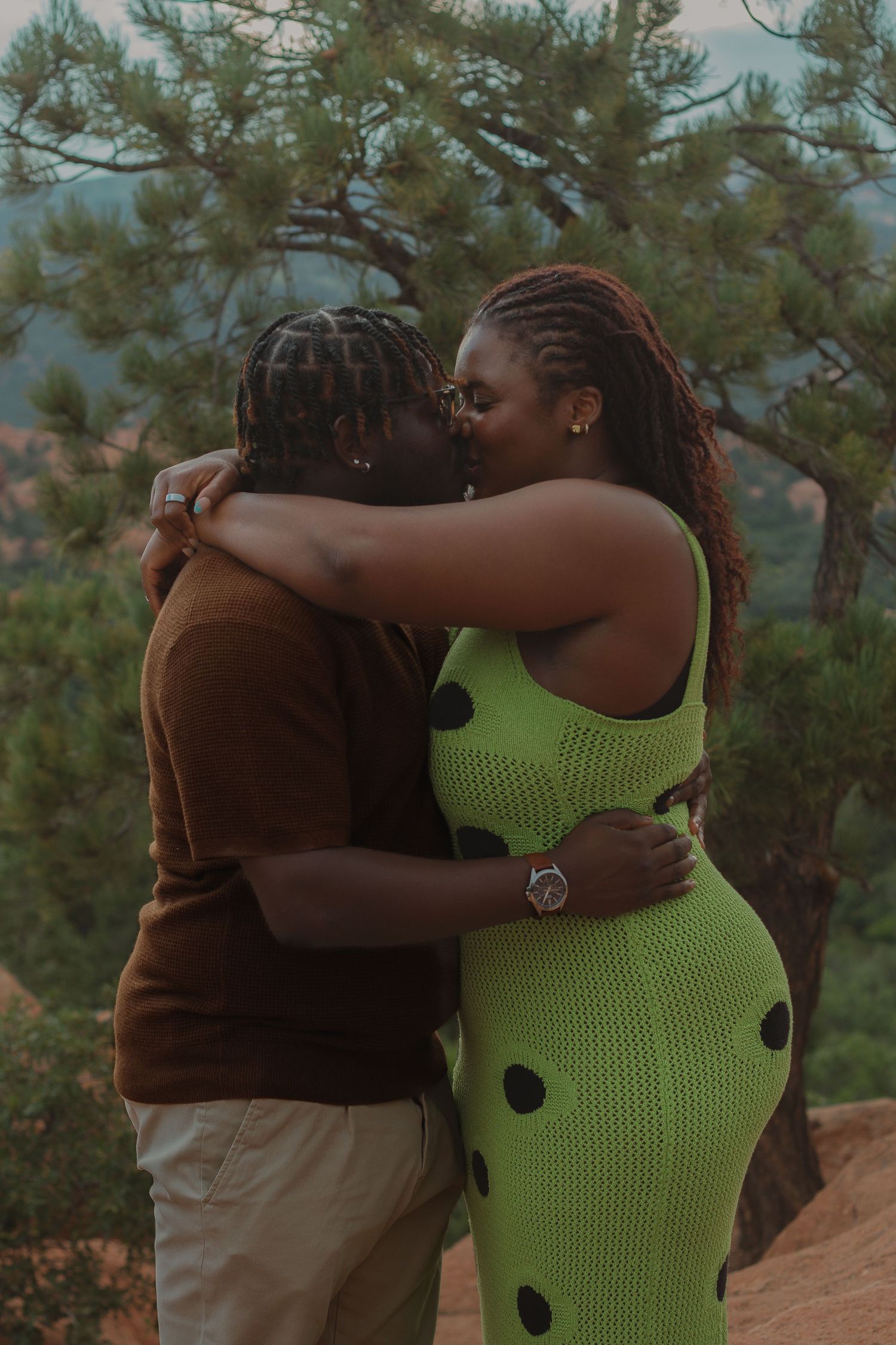 A romantic embrace between two people on a mountainside, with pine trees in the background.