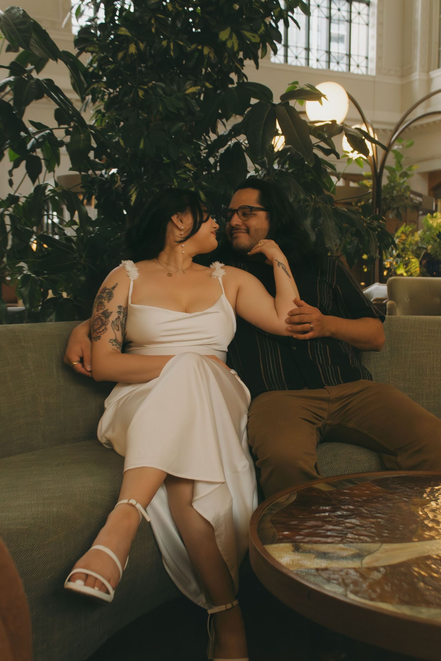 A romantic couple shares an intimate moment on a couch surrounded by greenery in an indoor setting.