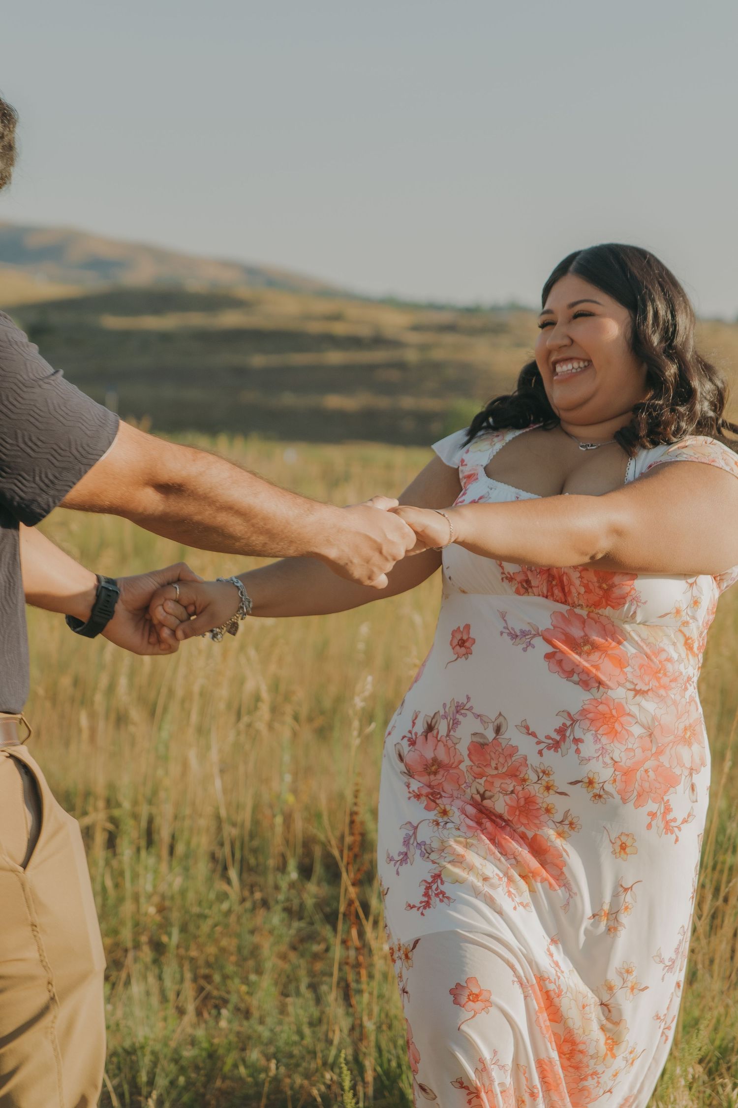 A couple dances and laughs together in a field during golden hour with hills in the background.