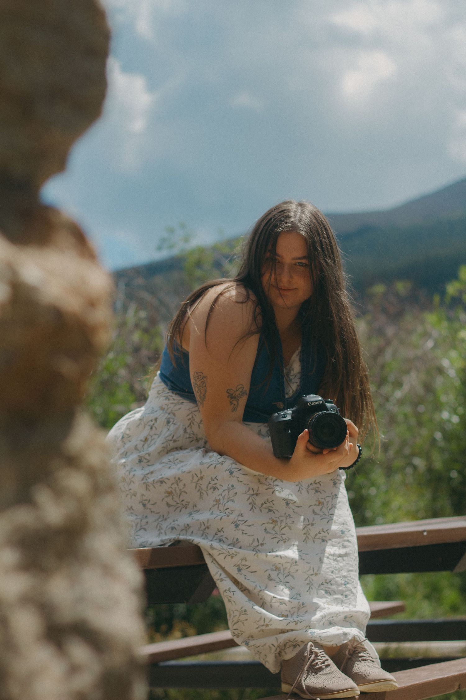 Photographer in white layered skirt and blue top holding camera sits on wooden railing with mountains in background.