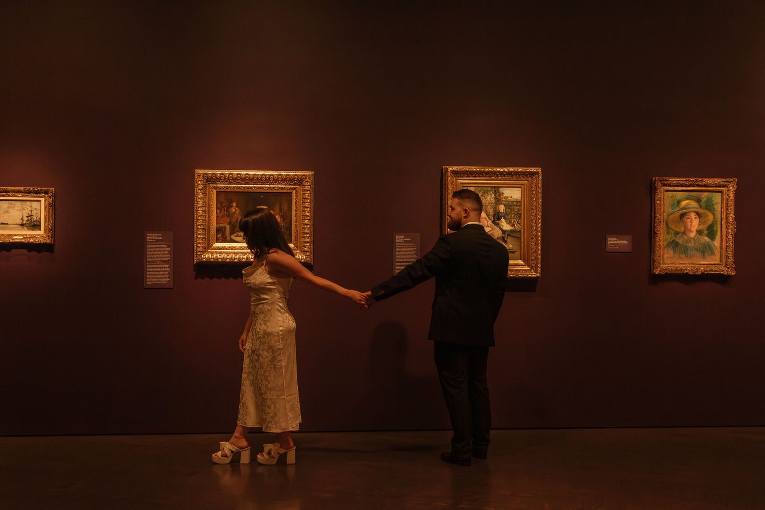 Art gallery interior with dark walls displaying framed artwork in warm lighting and visitors viewing the collection.
