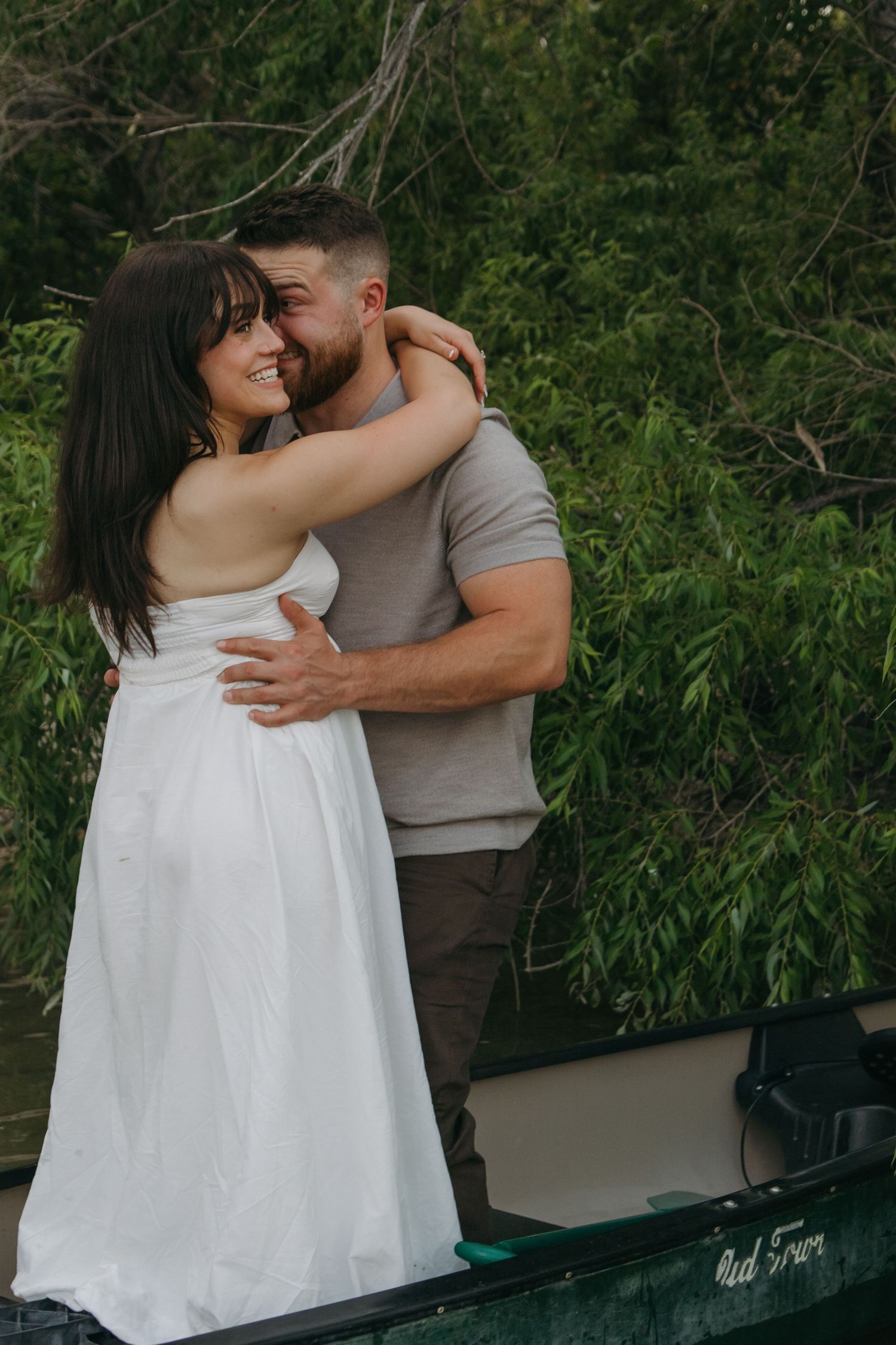 A couple embraces while standing next to a small boat near lush greenery.