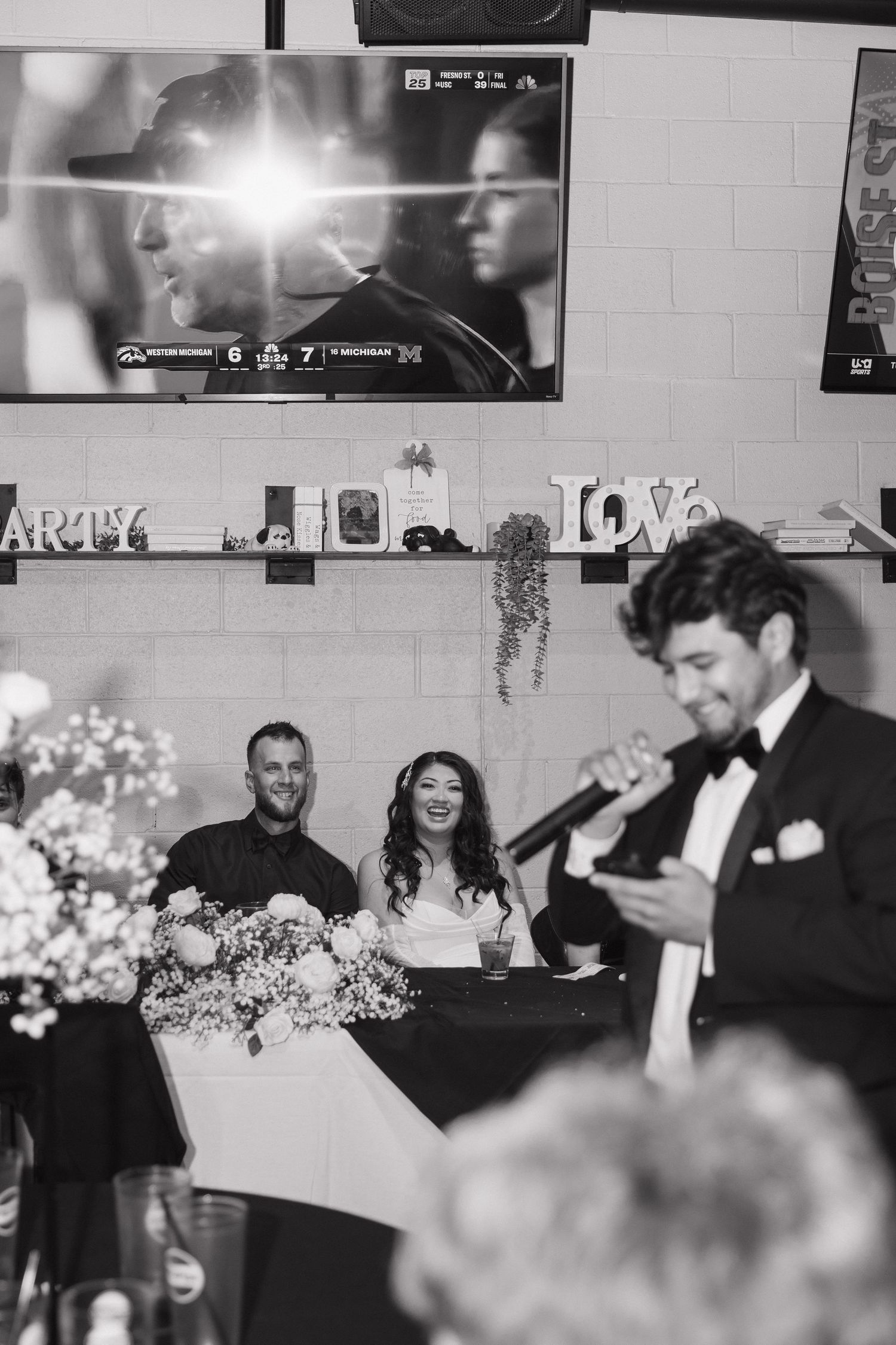 Black and white photo of groom giving speech while bride and groom smile at wedding reception.