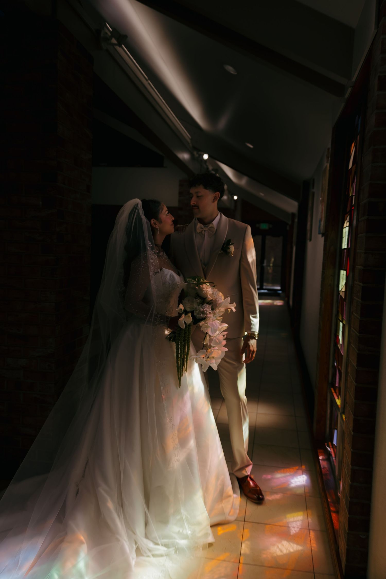 Bride and groom share a romantic moment in a dimly lit church hallway with stained glass light reflections on the floor.
