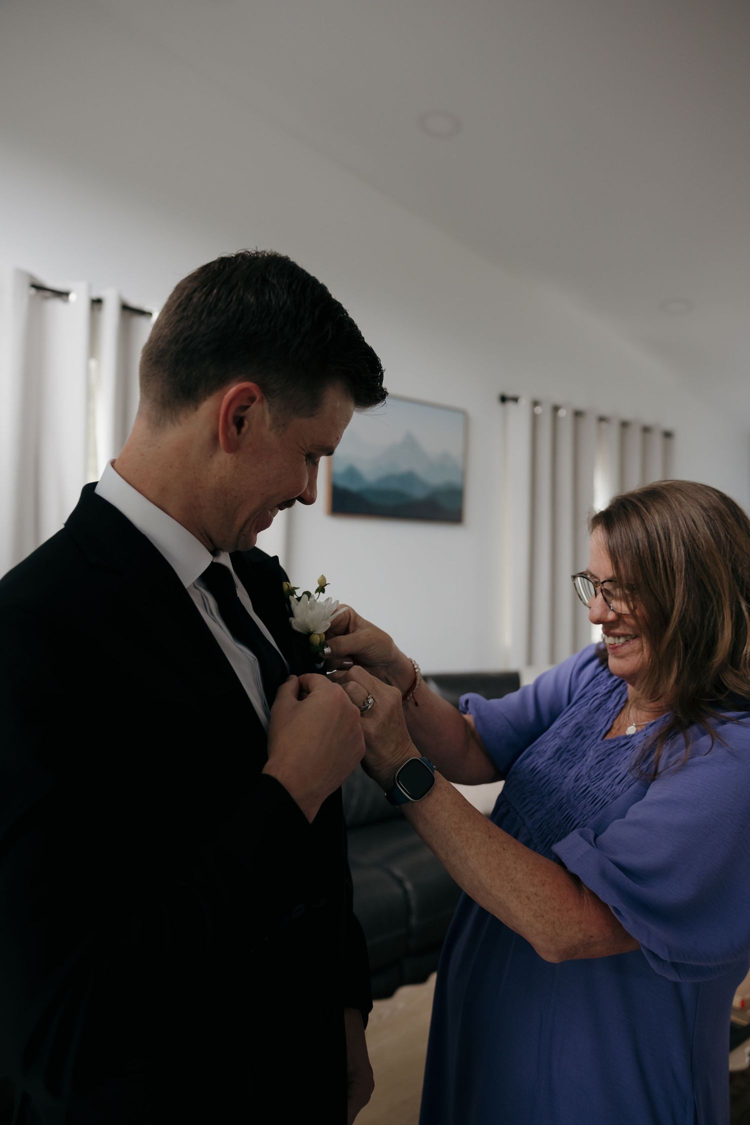 Woman in blue dress pinning a white boutonniere on a groom in a black suit before the wedding ceremony.