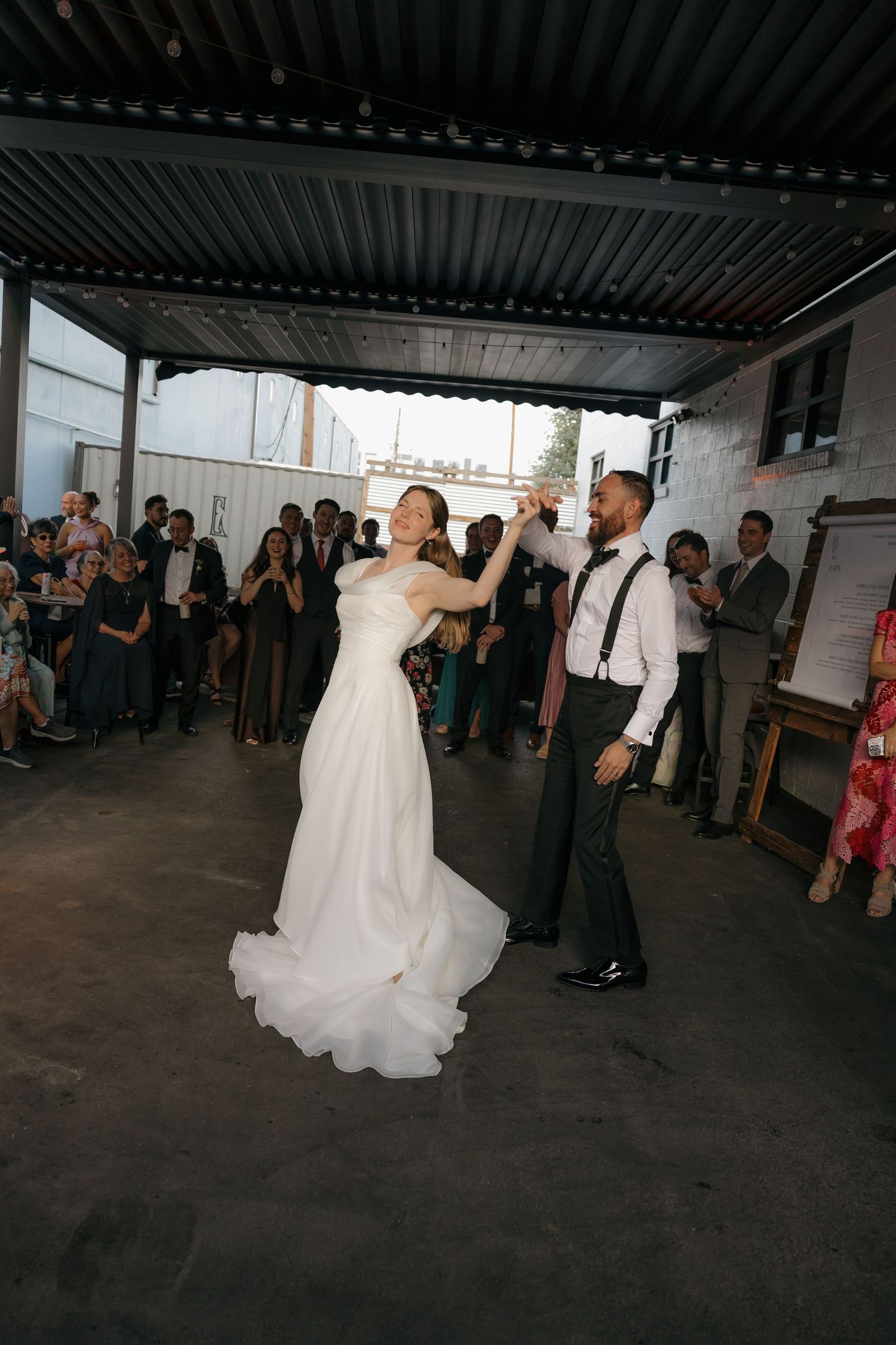 Bride and groom sharing first dance in outdoor industrial wedding venue surrounded by guests.