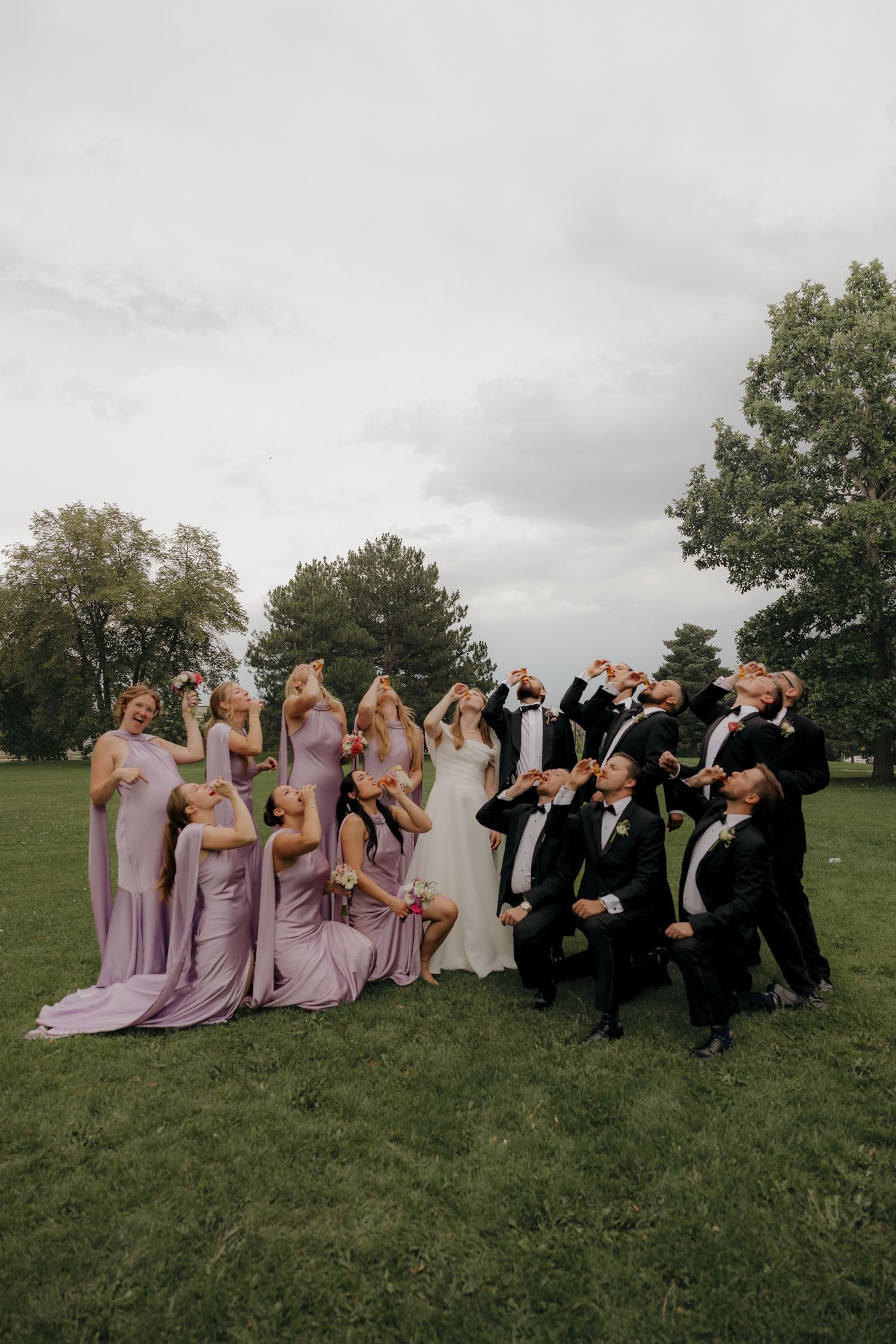 Wedding party in lavender bridesmaid dresses and black suits toasting drinks on a green lawn.
