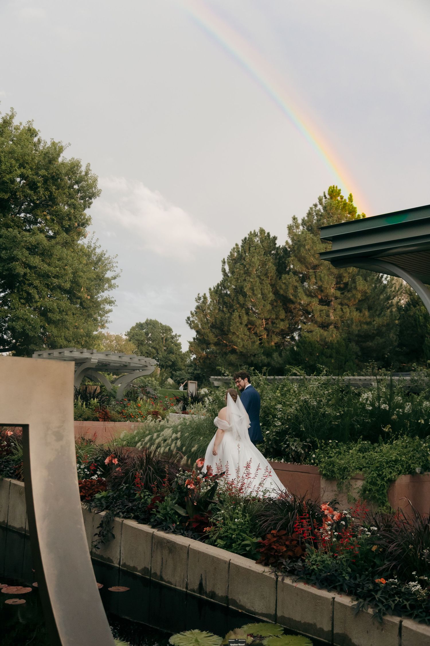 Bride and groom stand in a colorful garden with a stunning rainbow arching over lush trees on their wedding day.