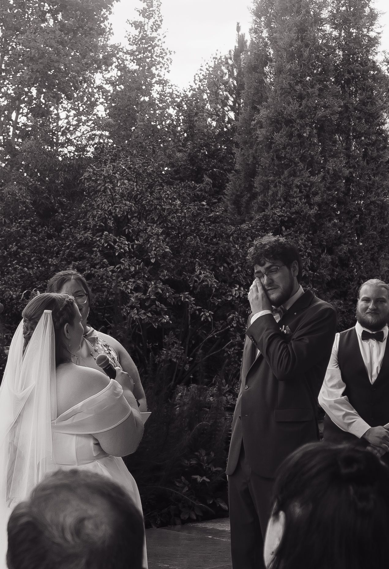Black and white photo of emotional bride and groom crying during outdoor wedding ceremony with guests watching.