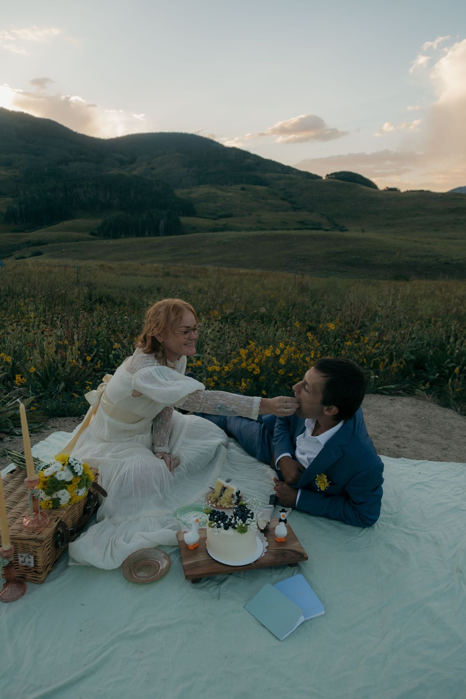 Bride feeding groom cake during outdoor mountain meadow picnic wedding celebration at sunset.