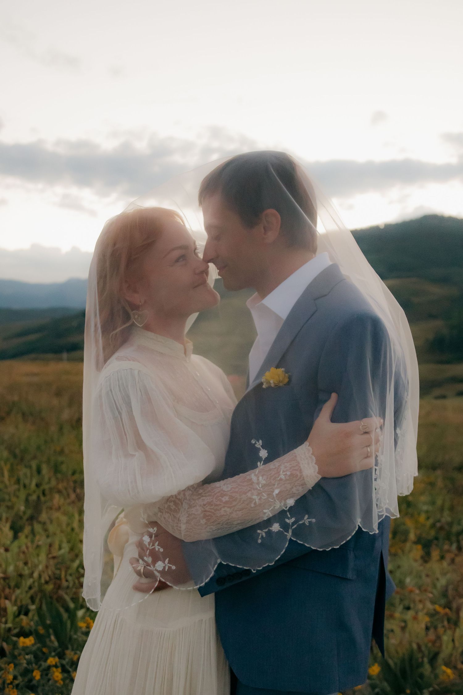 Bride and groom share intimate moment under flowing veil in mountain wildflower field at golden hour.