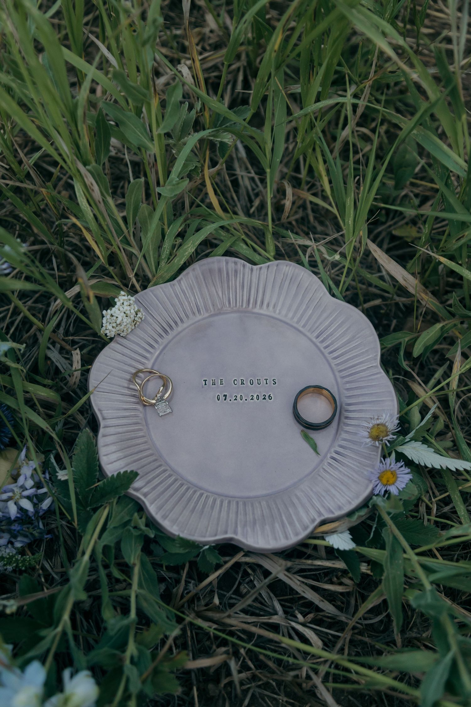 Ceramic ring dish with wedding bands resting in grass with wildflowers.