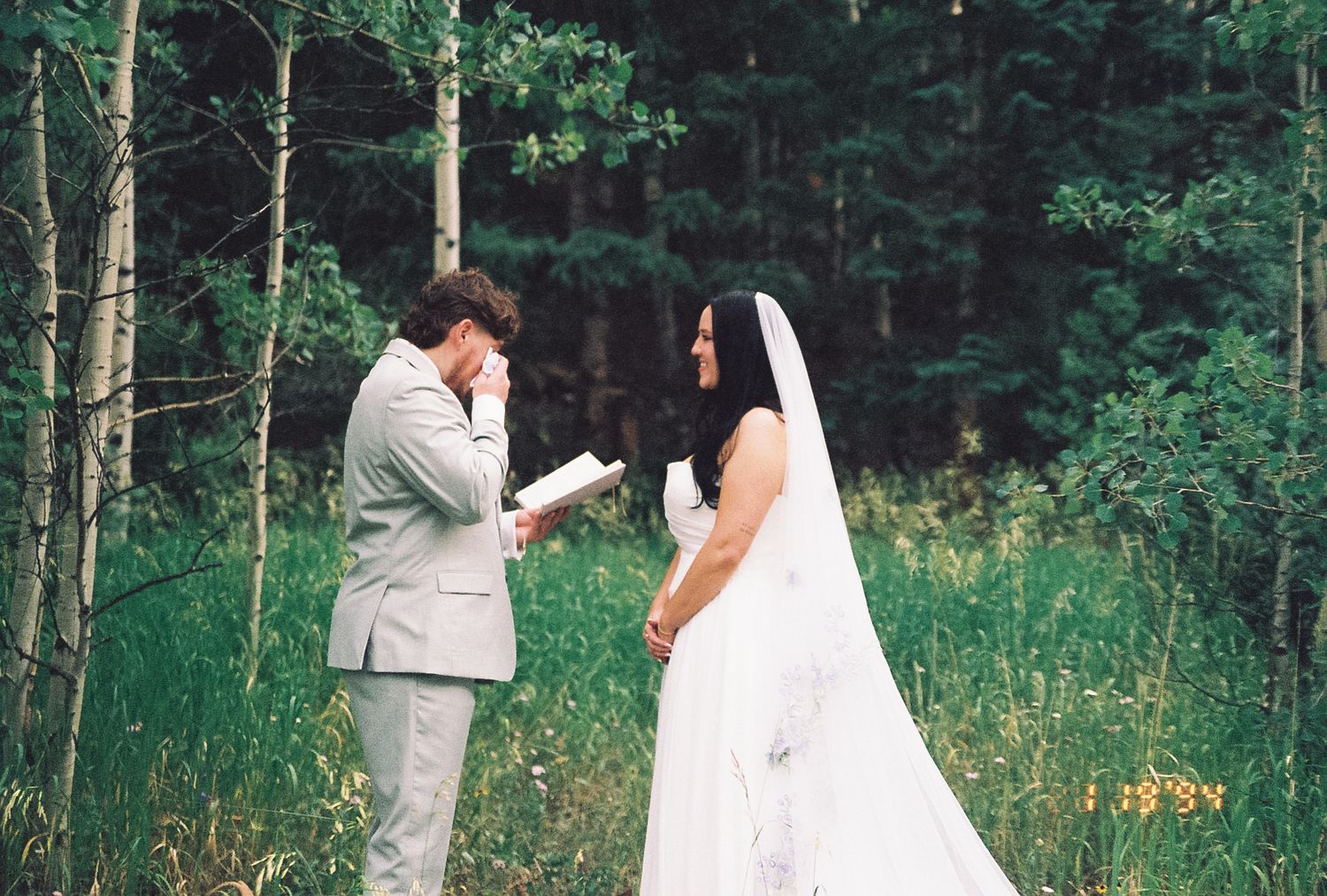 Emotional groom wipes tears while reading vows to his bride in a white gown in a forest setting.