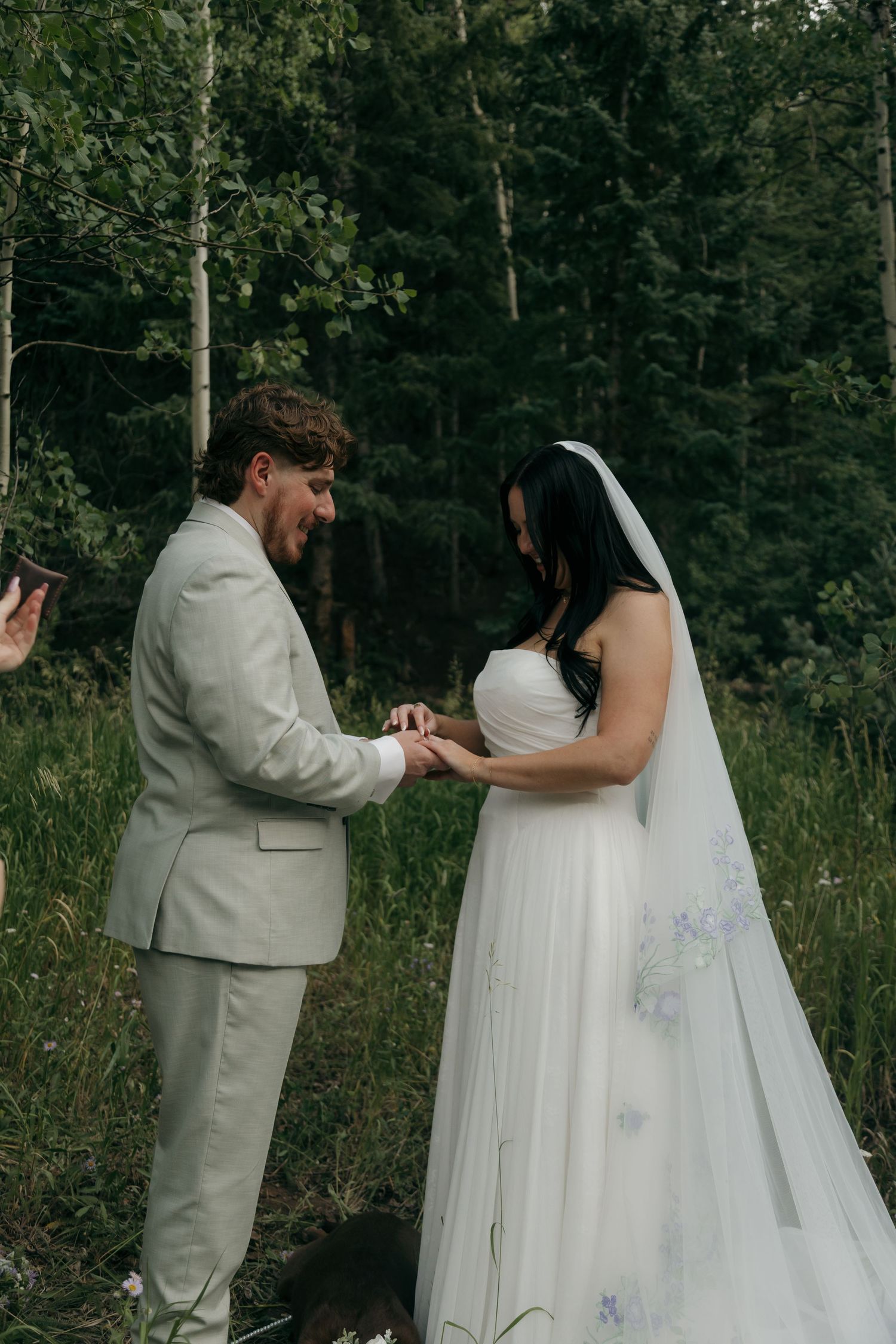 Groom places wedding ring on bride's finger during intimate forest elopement ceremony in summer woods.