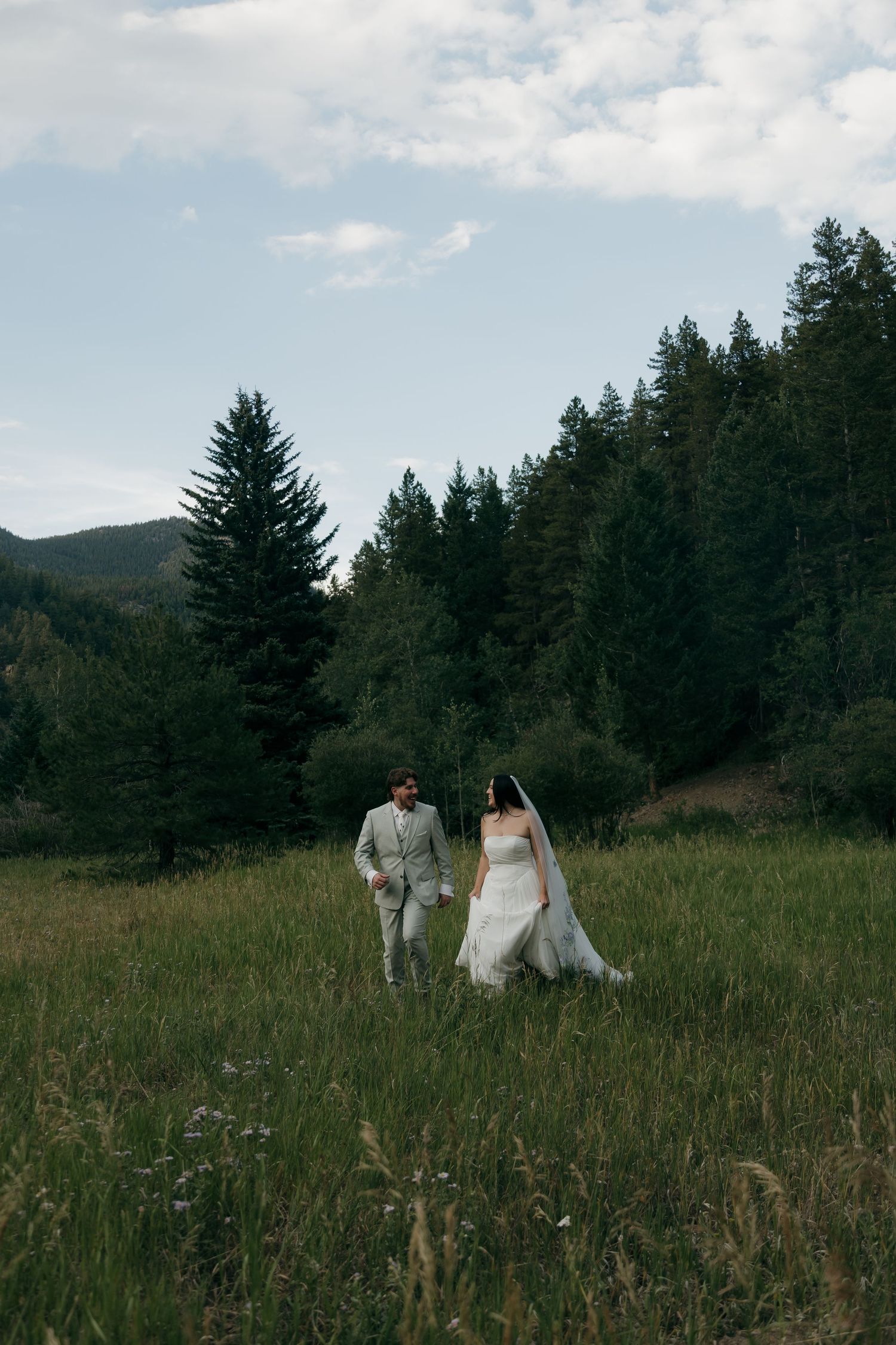 Bride in white gown with veil and groom in gray suit walk hand-in-hand through a lush mountain meadow.