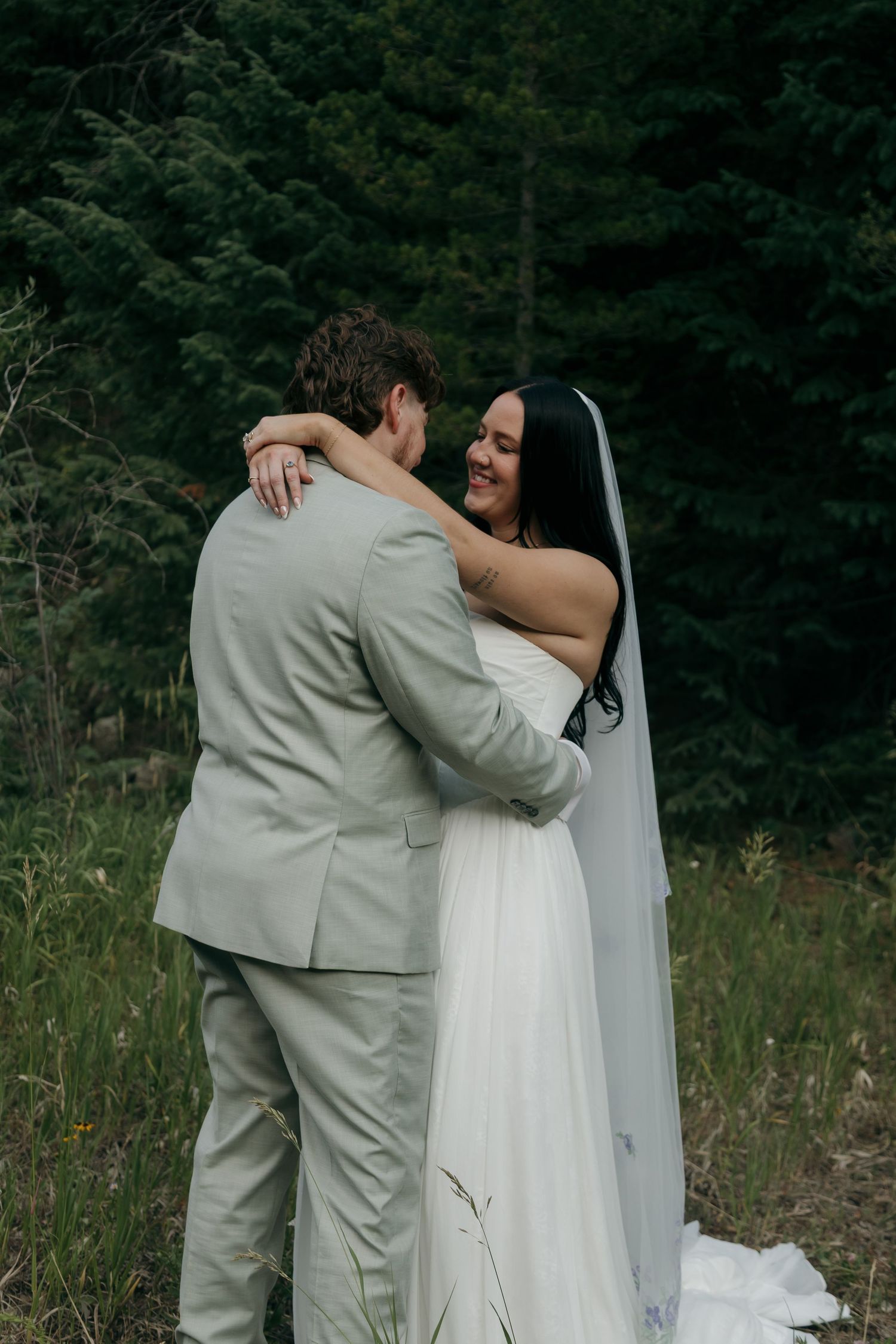Smiling bride in white wedding dress embraces groom in light gray suit amid a lush forest backdrop during outdoor celebration.