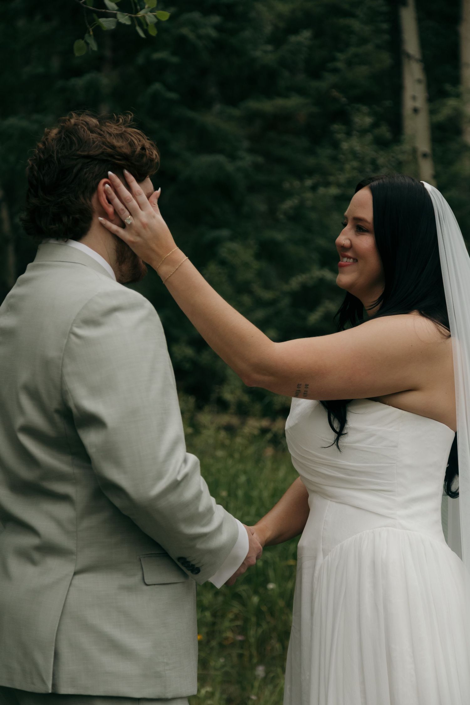 Bride in white gown and veil tenderly touches grooms face during outdoor wedding ceremony in forest.