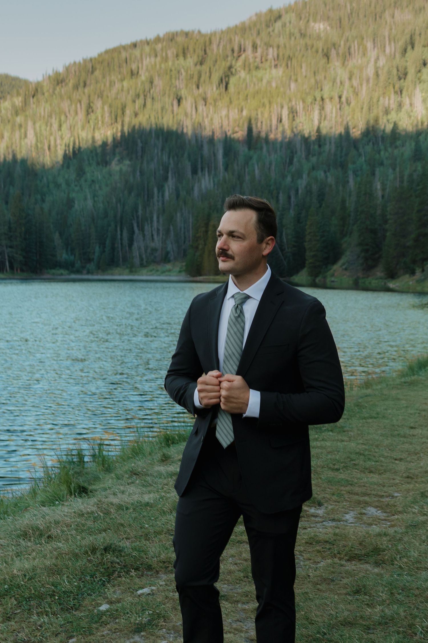 Groom in black suit poses by mountain lake with evergreen forest in background.