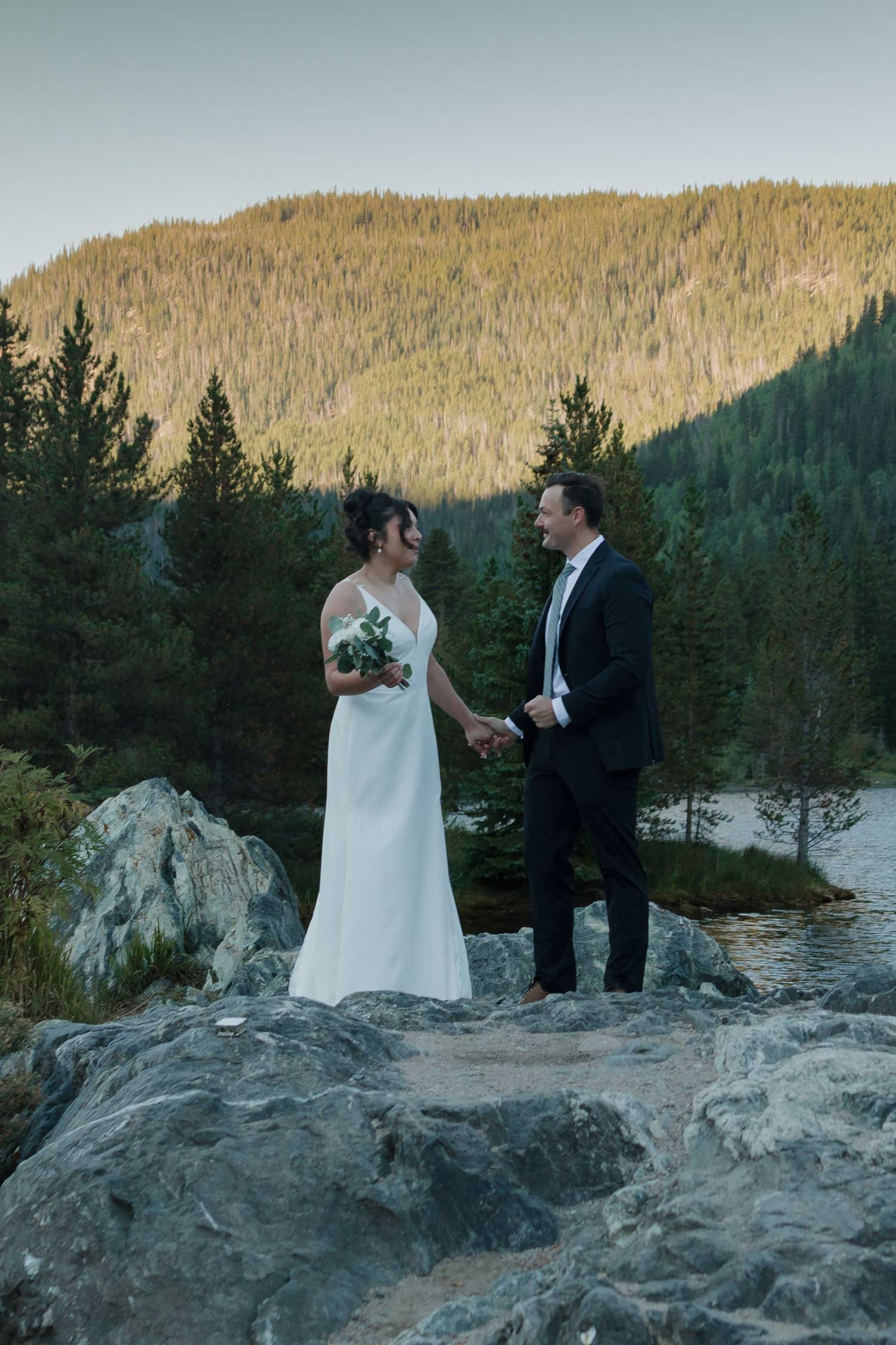 A wedding couple shares an intimate moment on rocky terrain overlooking a serene lake at sunset.