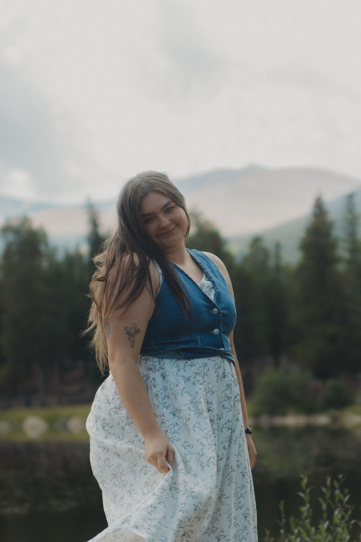 A person in a white floral dress and blue vest dances joyfully by a serene lake with mountains in the background.