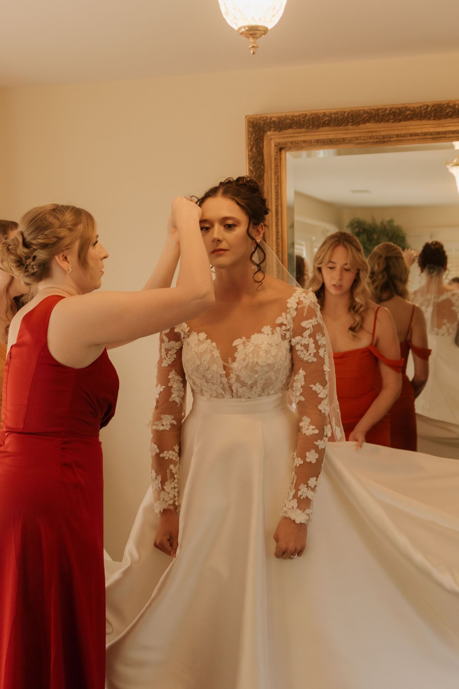 Group preparing bride's wedding dress in room with bridesmaids in red dresses.