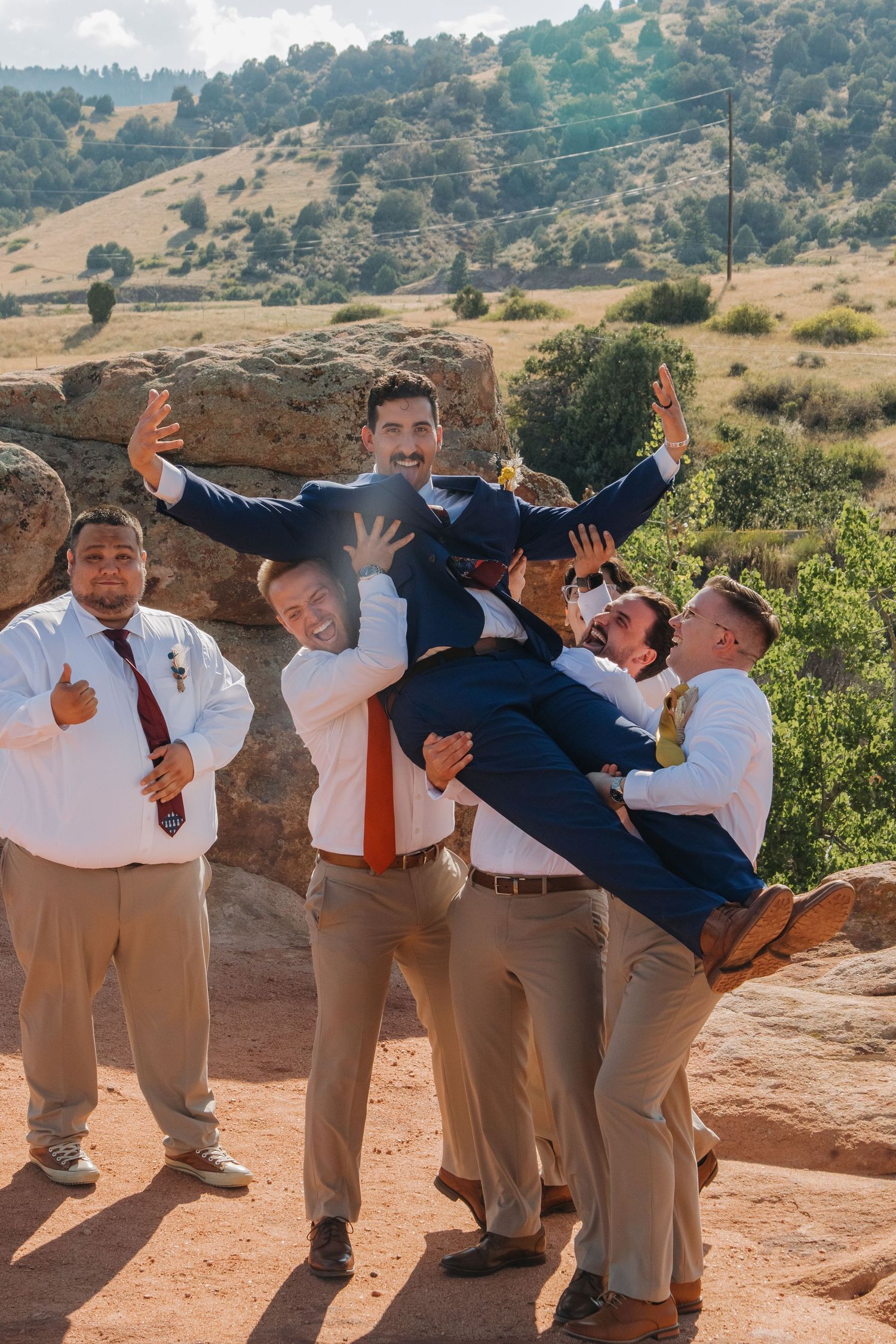 Groomsmen in matching attire strike playful jumping poses on red rocks against a desert landscape backdrop.