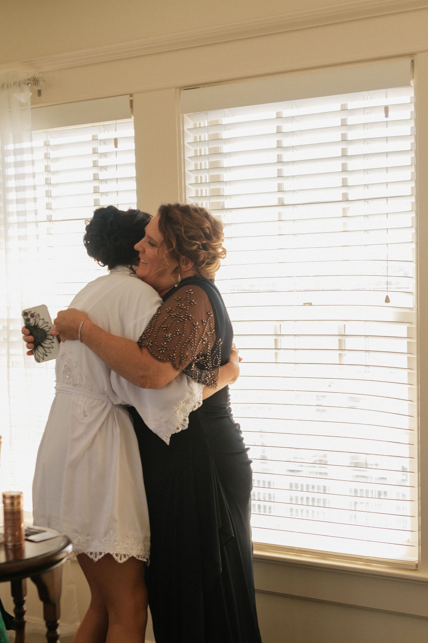People embracing in a sun-lit room with white blinds.