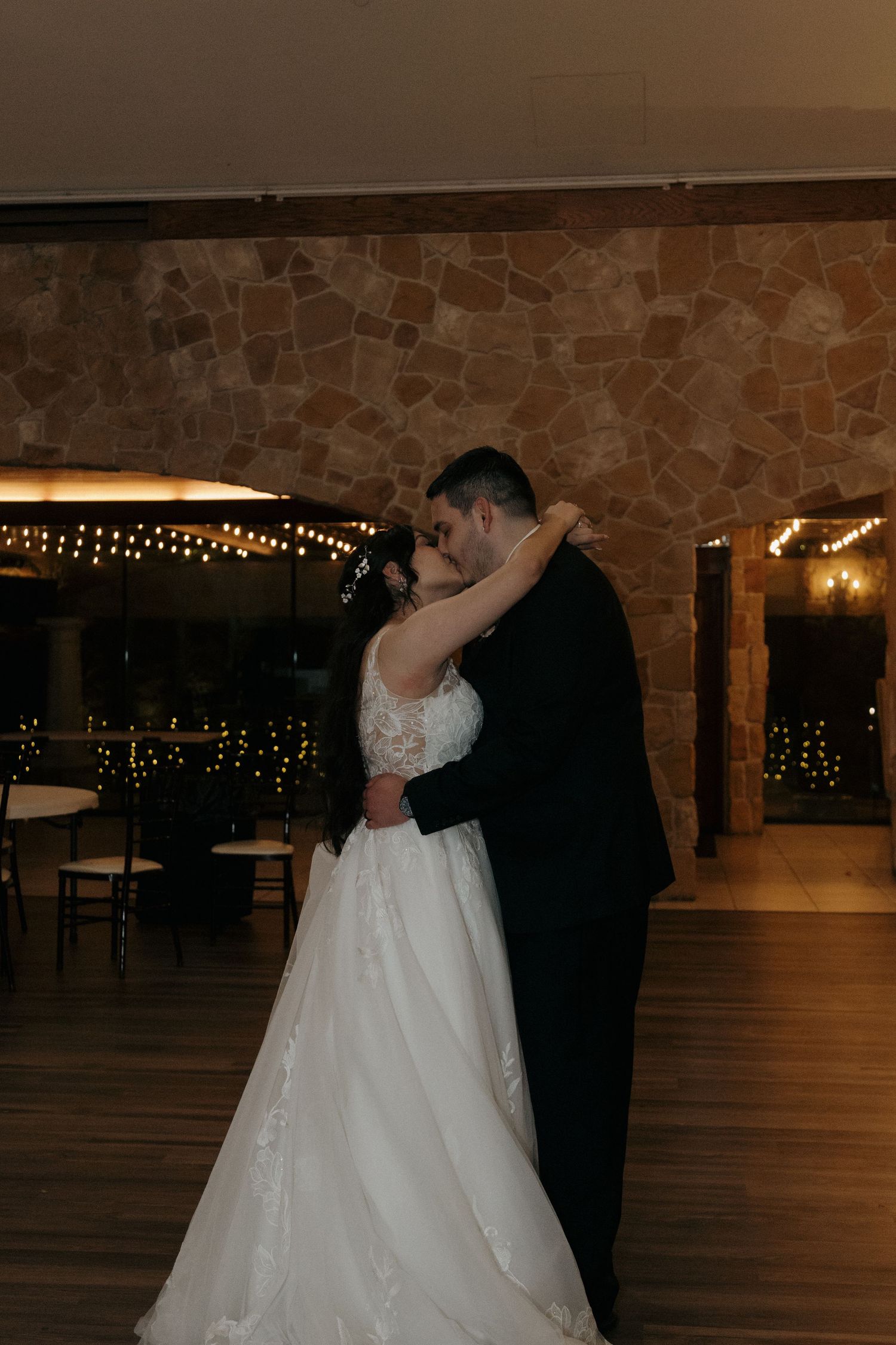 Bride and groom share a romantic first dance embrace in a warmly lit rustic stone wedding venue.