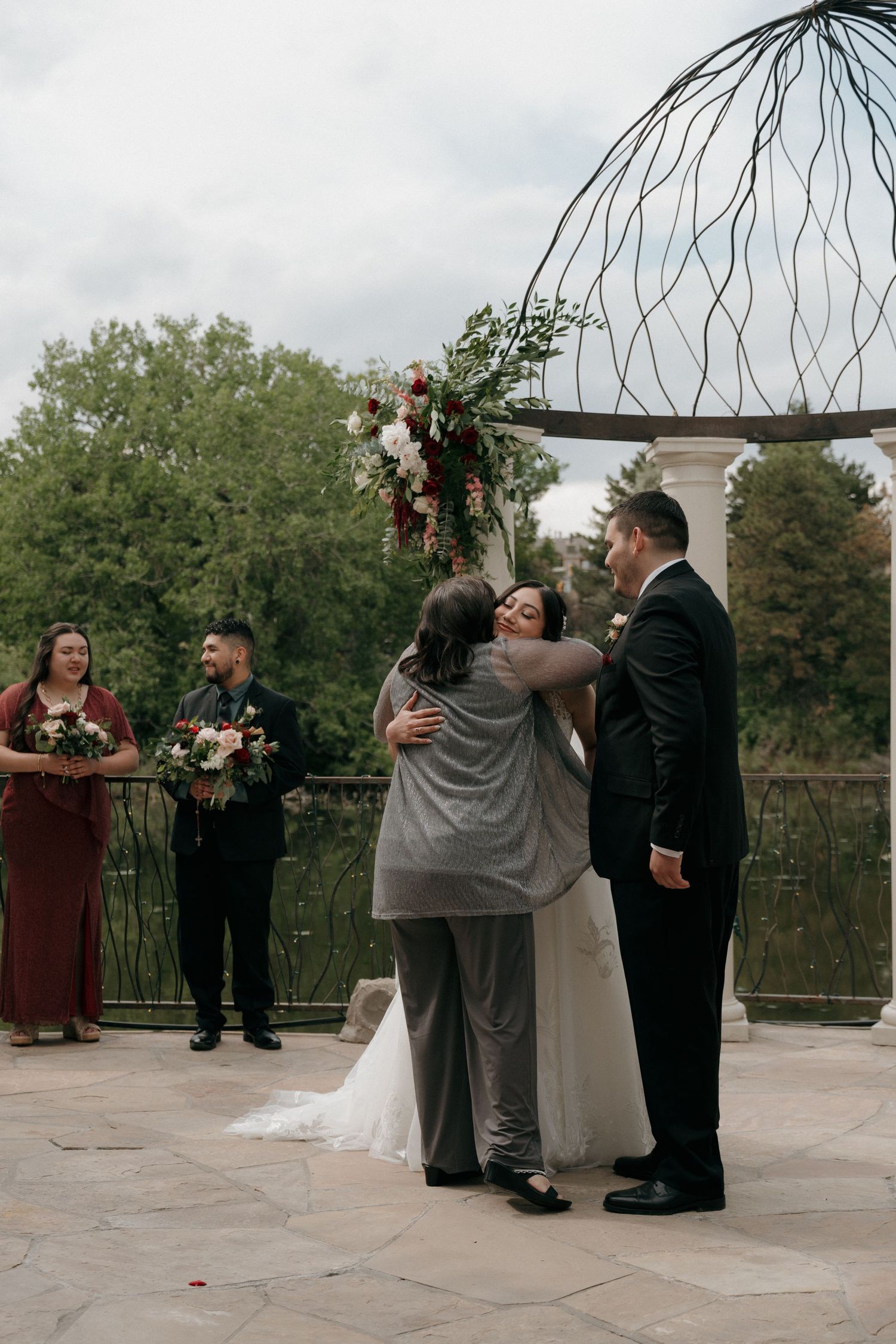 A bride shares a warm hug with a guest at an outdoor wedding ceremony beneath a decorative metal arch.